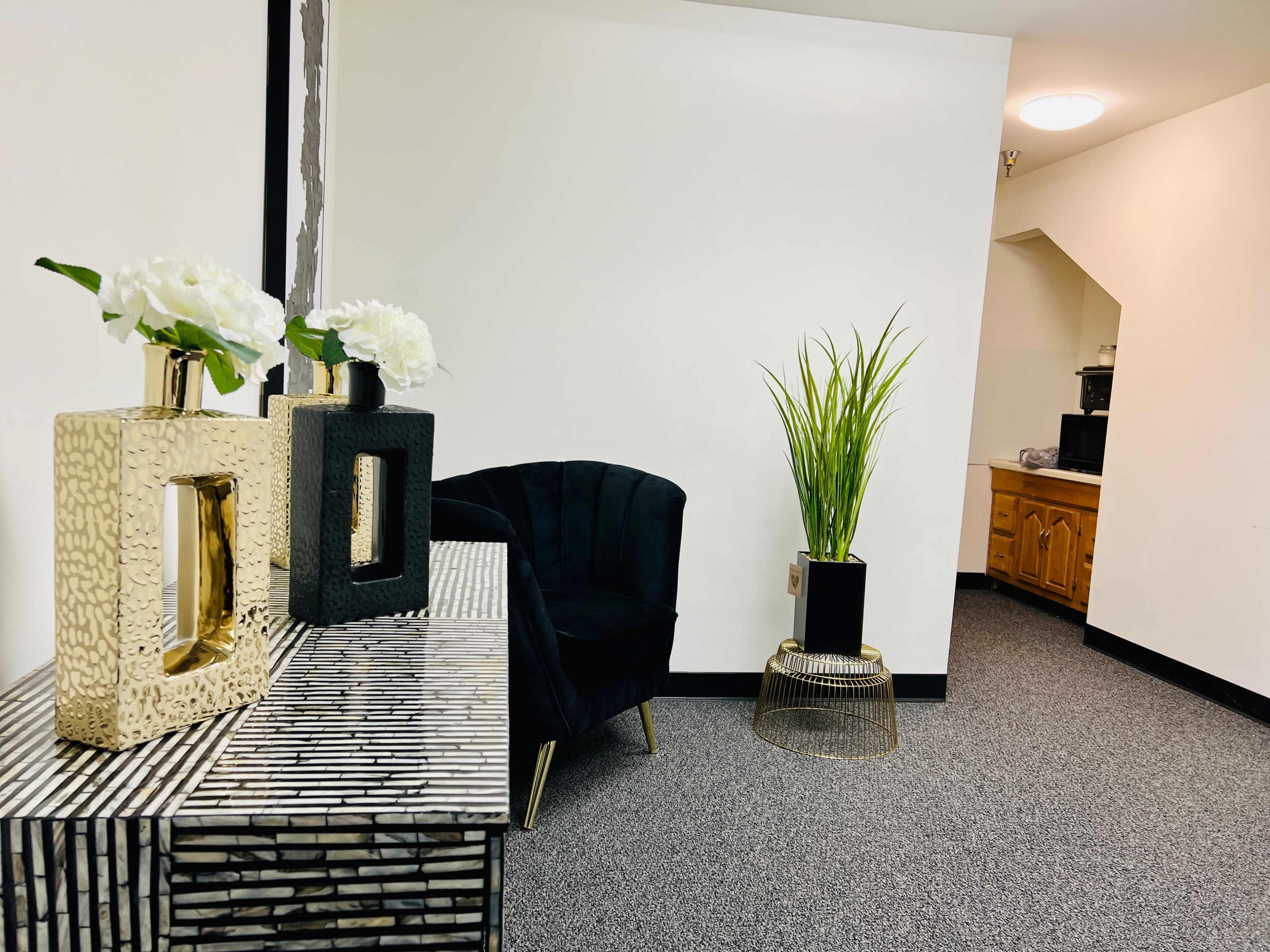 A small room featuring a patterned console table with decorative vases, a black upholstered chair, and a potted plant in a corner.