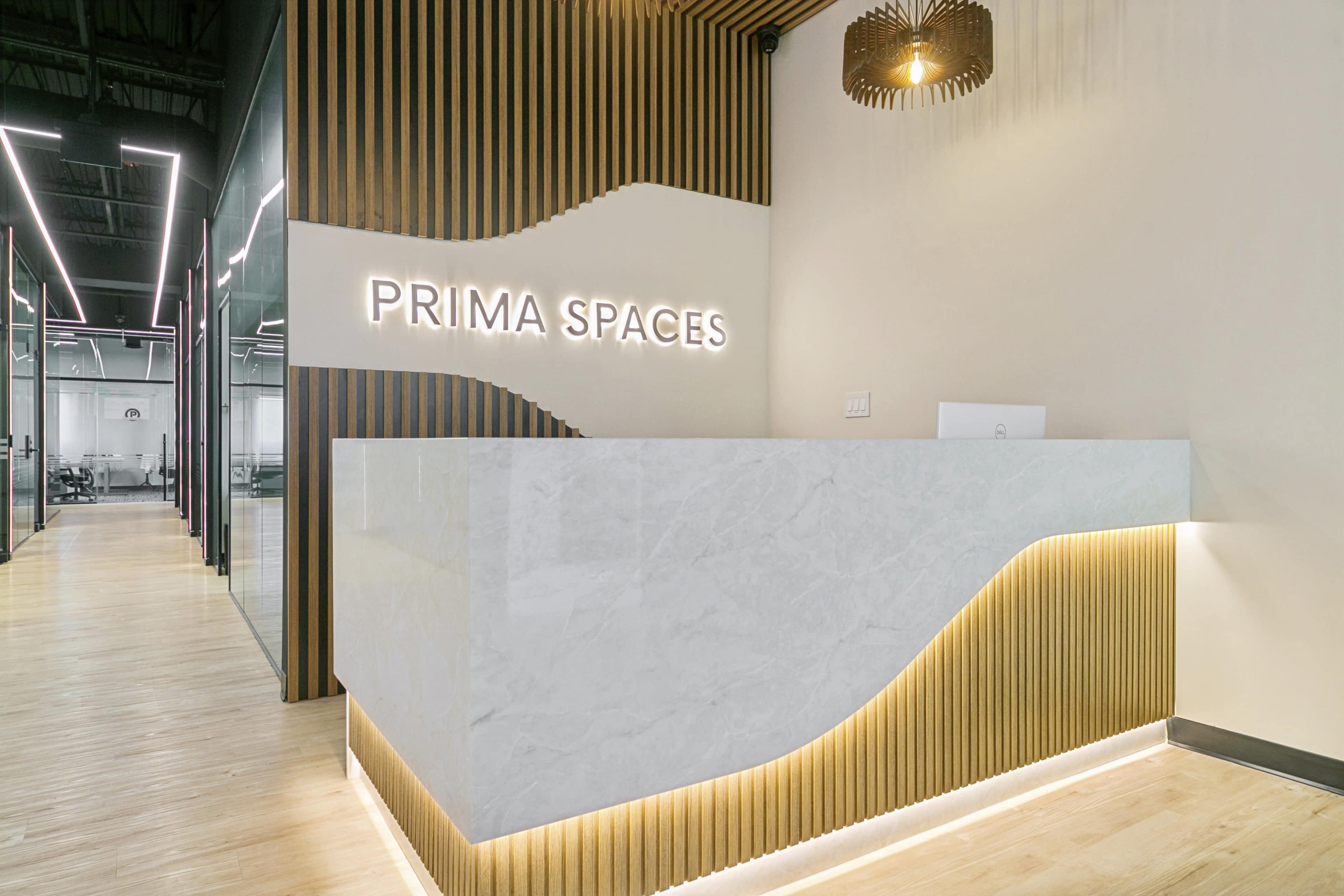 The image shows a modern reception area with a backlit marble desk and a wooden slat wall featuring the name "PRIMA SPACES."