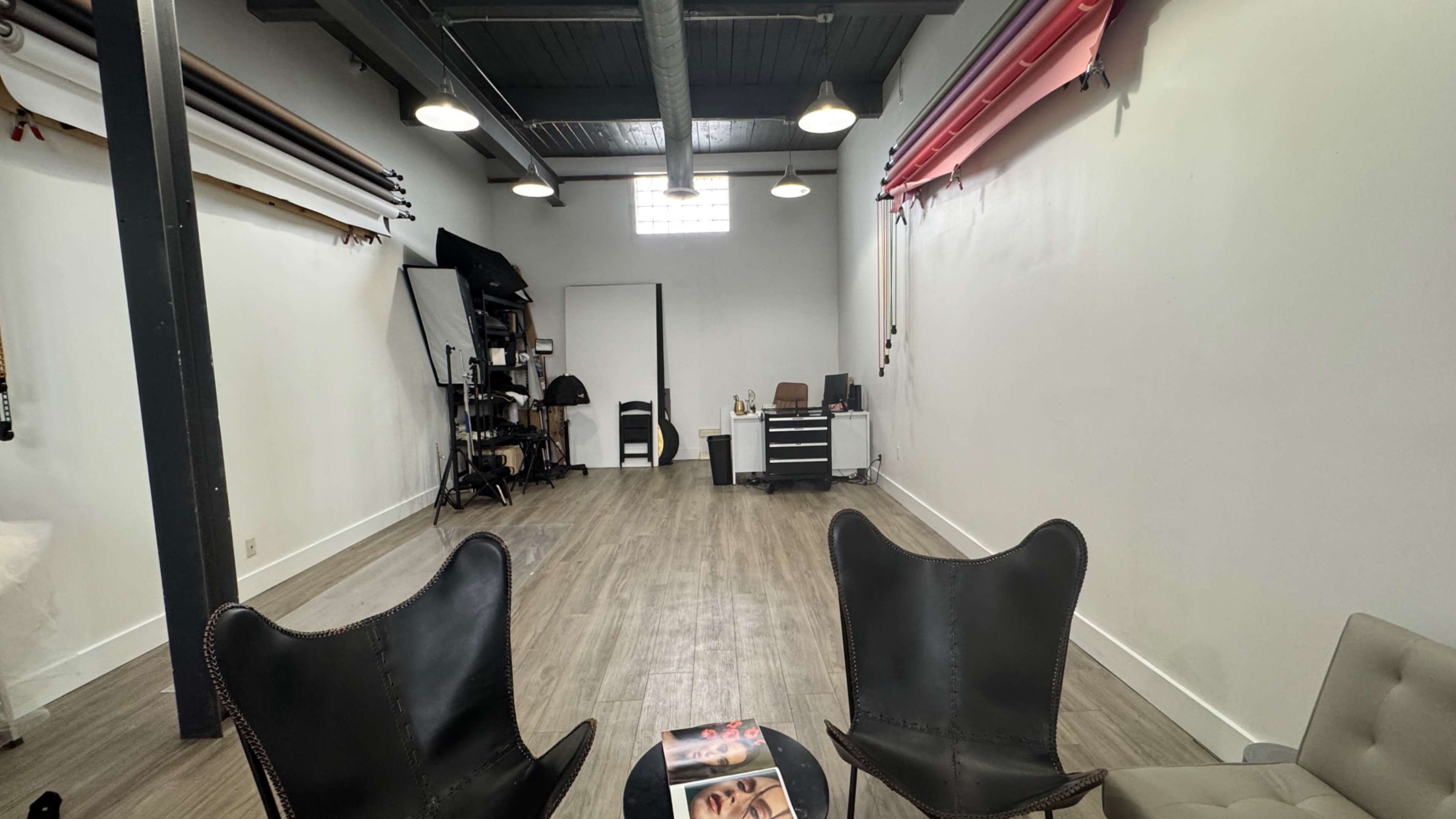 The image shows a spacious studio with two leather chairs facing a coffee table, while various photography equipment and backdrops are arranged along the walls.