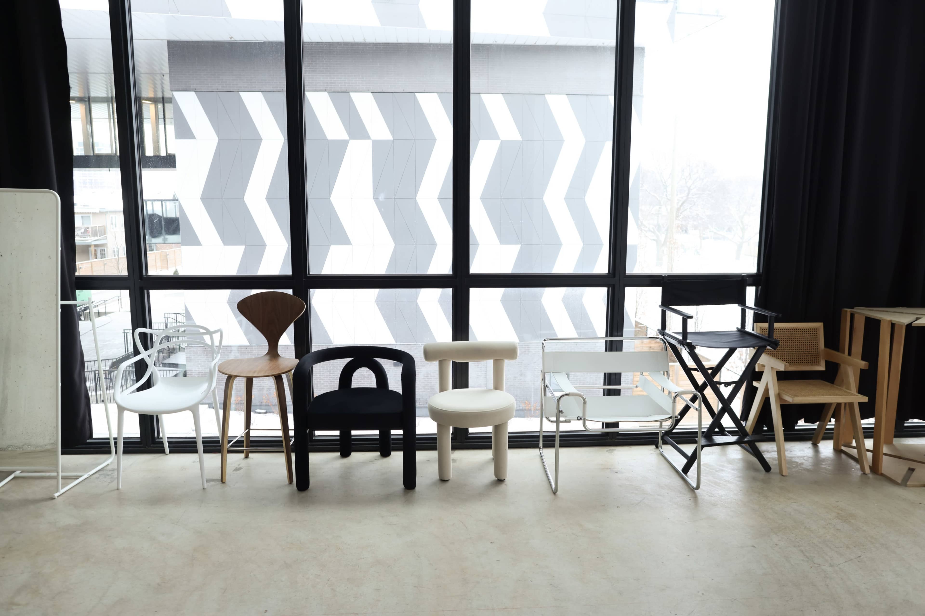 A row of various modern chairs is displayed in front of large windows, showcasing a geometric building pattern outside.