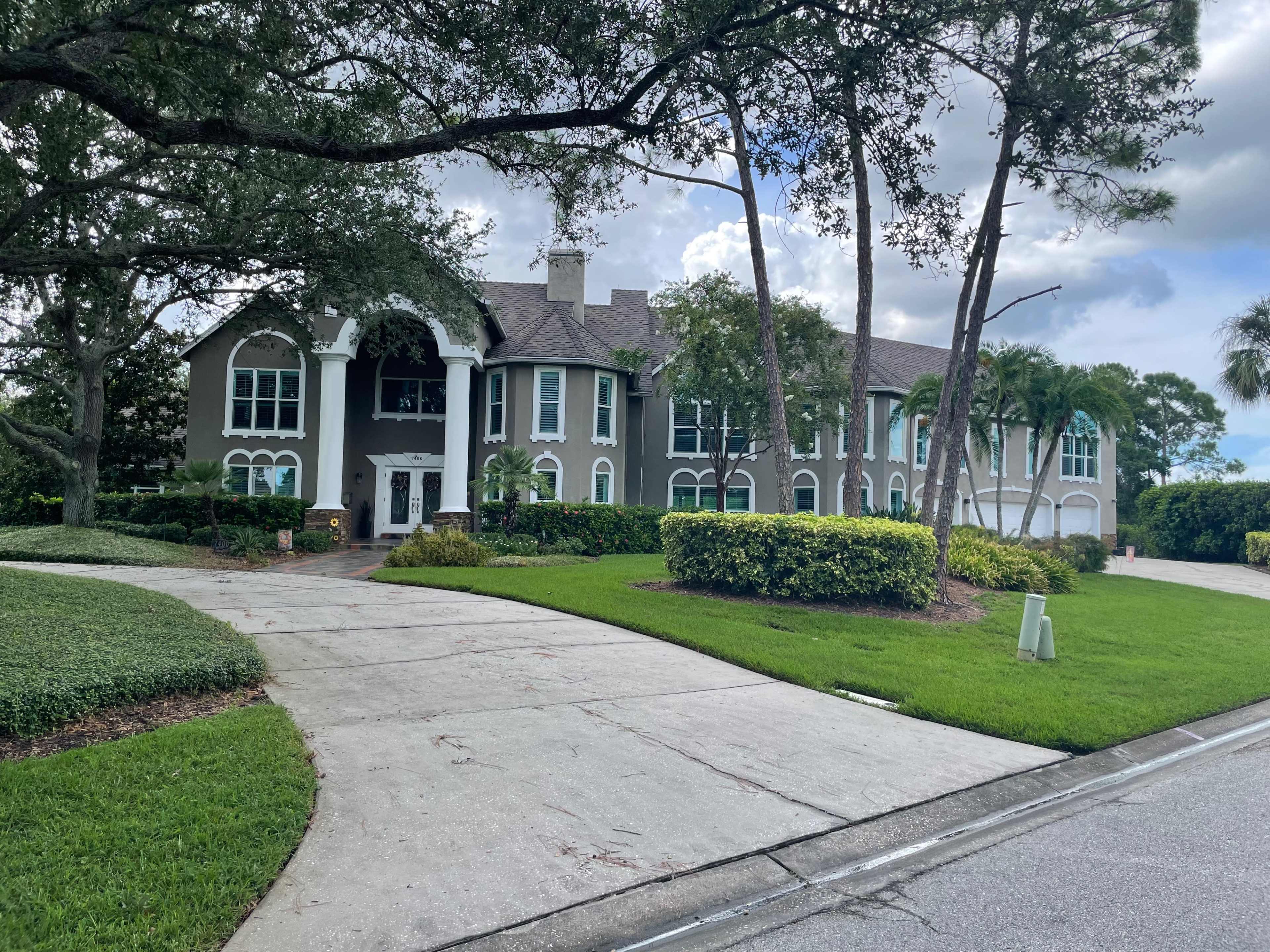 A large two-story house with a gray facade, multiple windows, and a wide driveway is surrounded by landscaped greenery.
