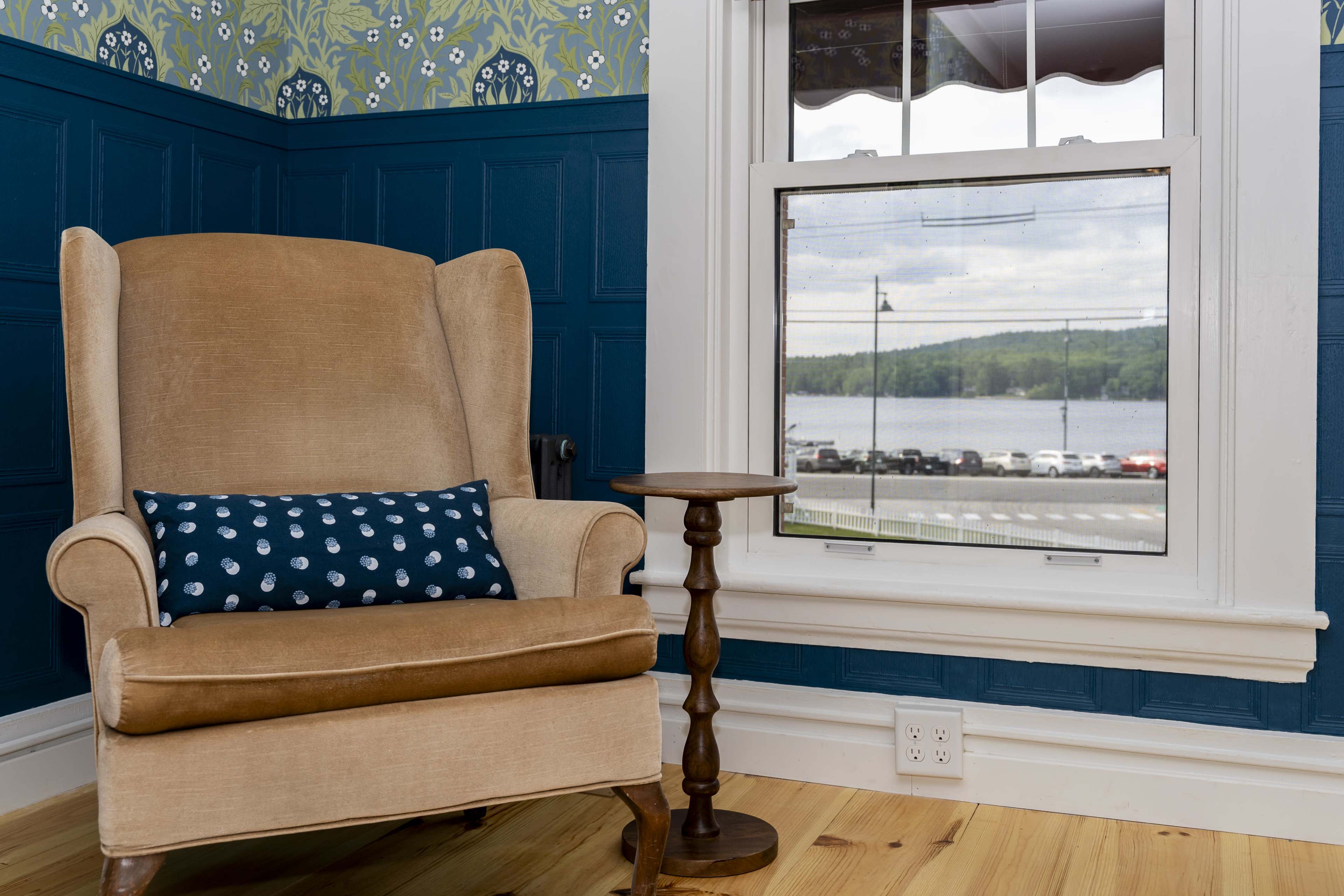 A beige armchair with a decorative blue pillow sits beside a wooden side table in a room featuring blue-painted walls and patterned wallpaper.
