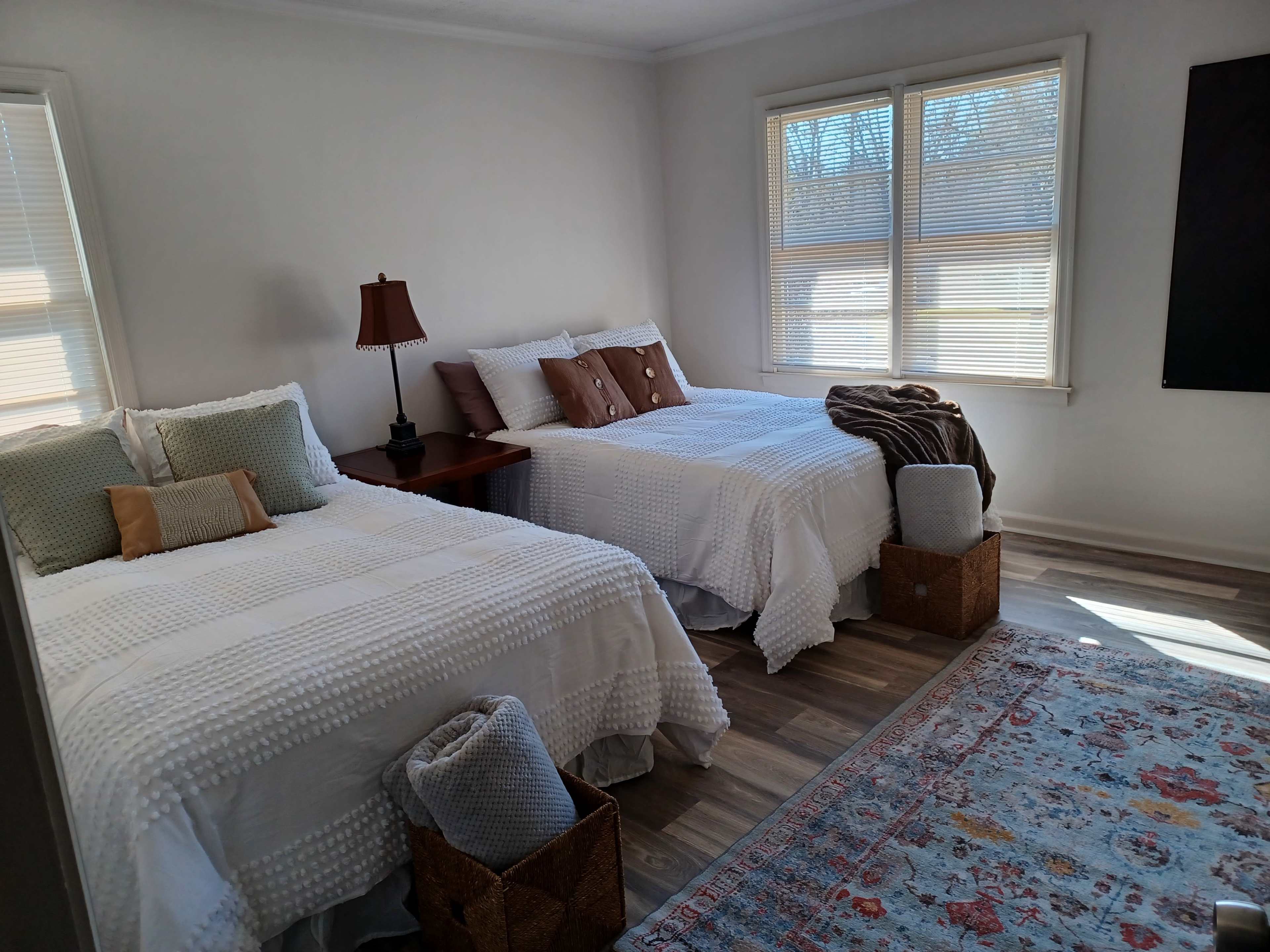 The image shows a bright bedroom with two separate beds dressed in white bedding, a small table with a lamp beside one bed, and a colorful rug on the floor.