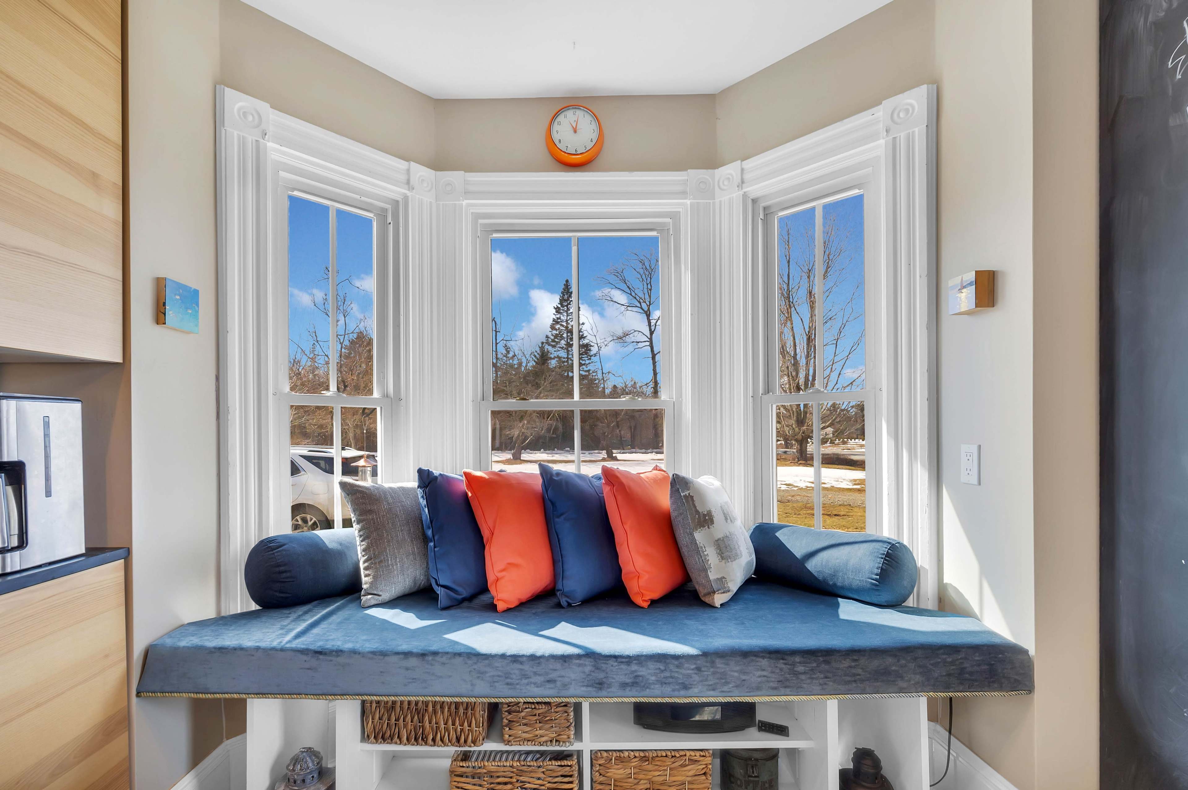 A cozy window nook with a blue cushion and decorative pillows is surrounded by large windows, providing a view of the outdoors.