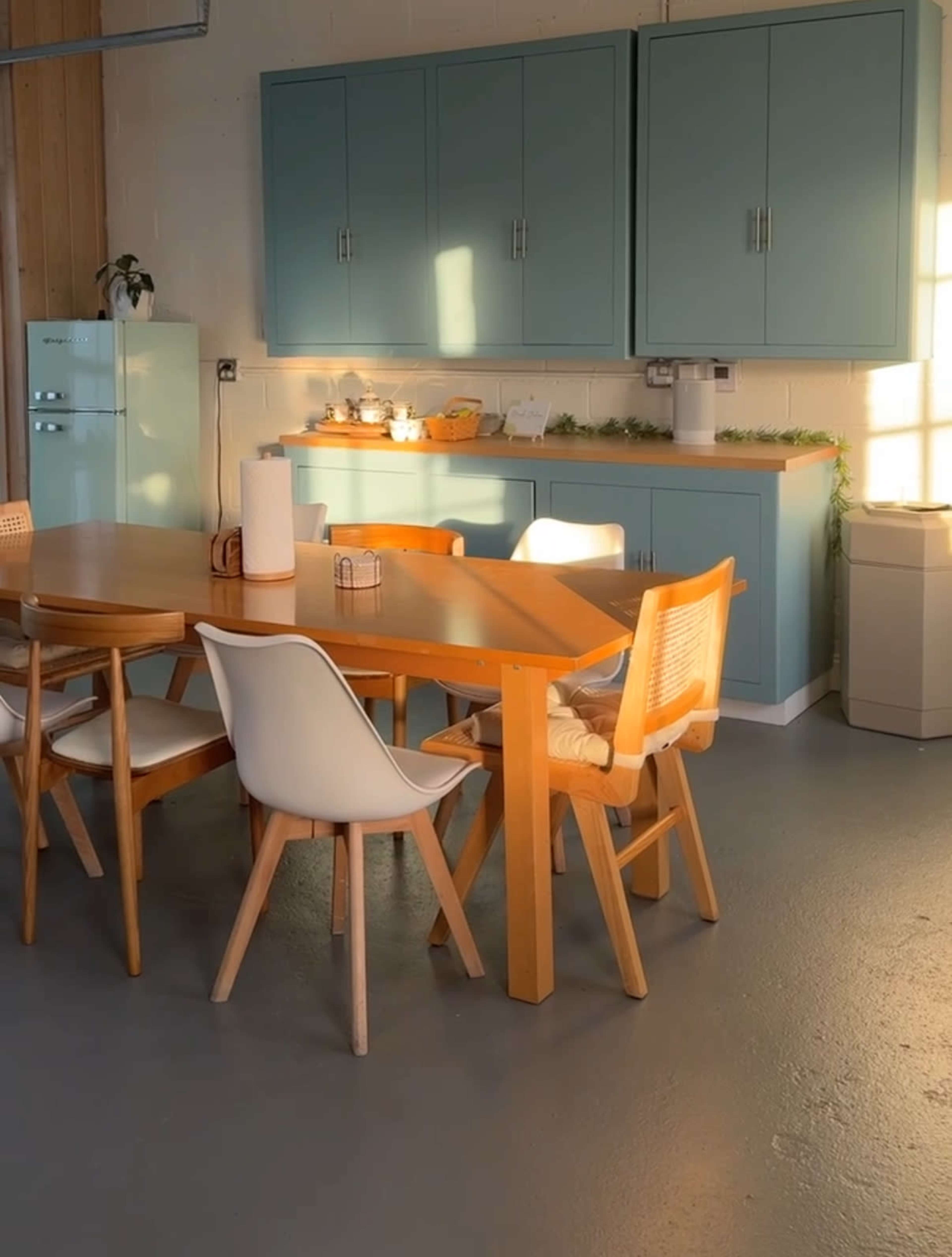 A wooden dining table with several chairs is positioned in a well-lit kitchen featuring blue cabinets and a retro refrigerator.