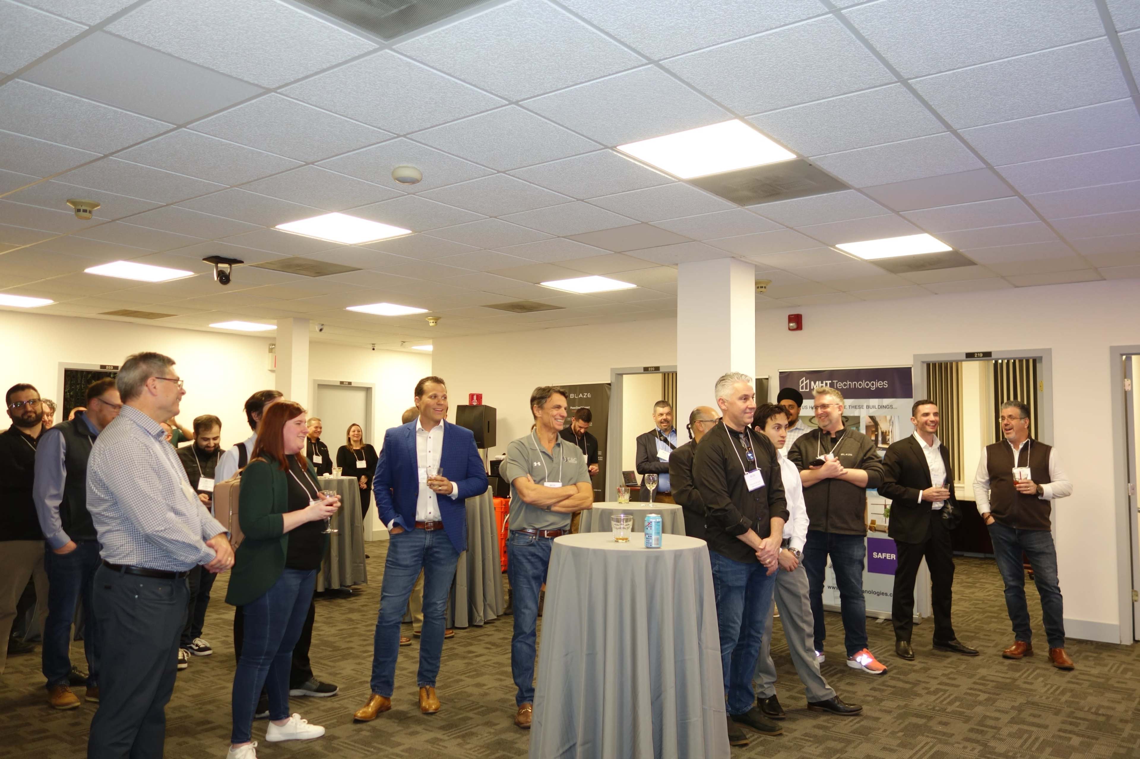 A group of attendees gathers in a conference room, listening to a speaker while holding drinks and standing around a few high tables.
