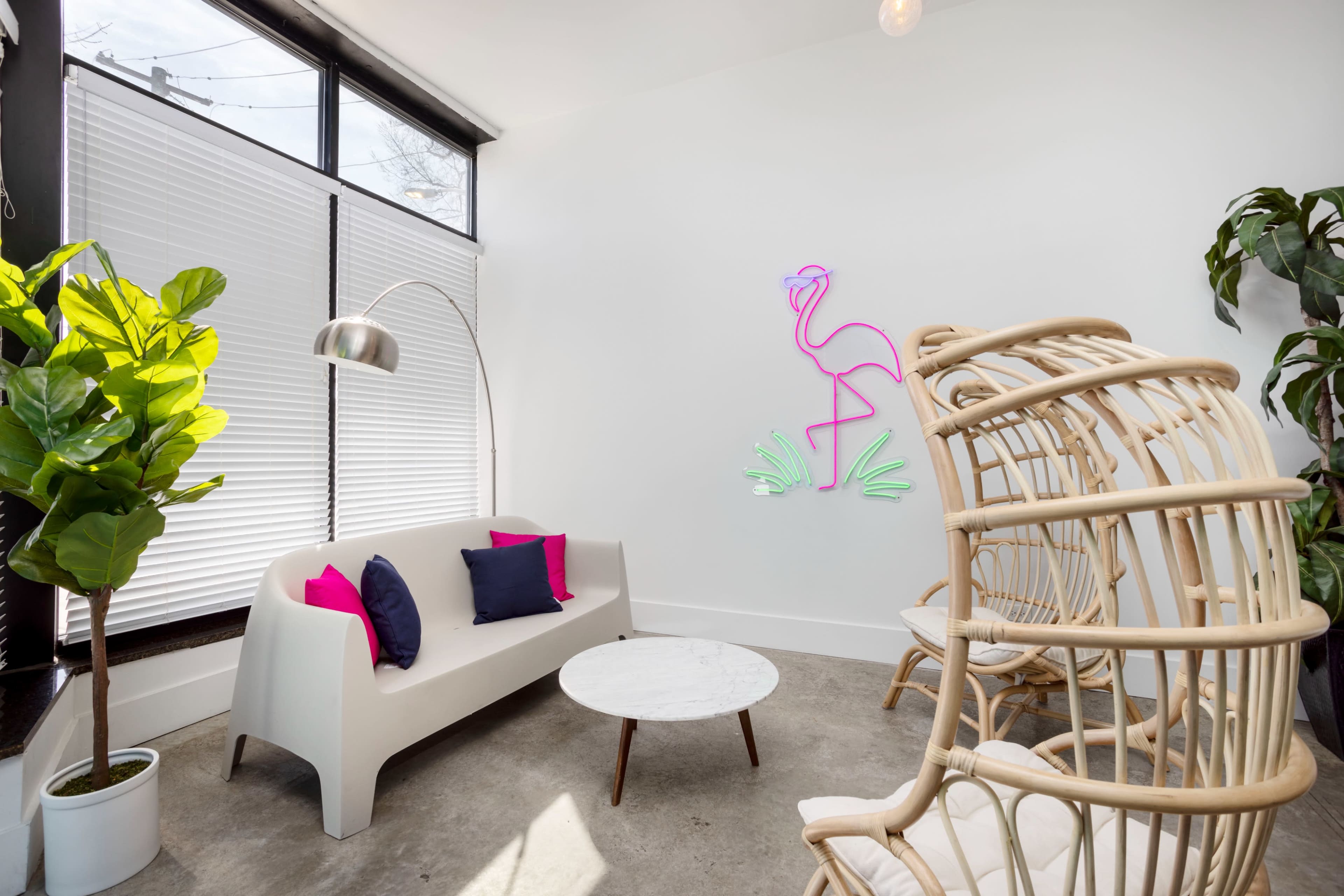 A modern seating area features a white sofa and a round coffee table, surrounded by large windows and decorative plants, with a neon flamingo art piece on the wall.