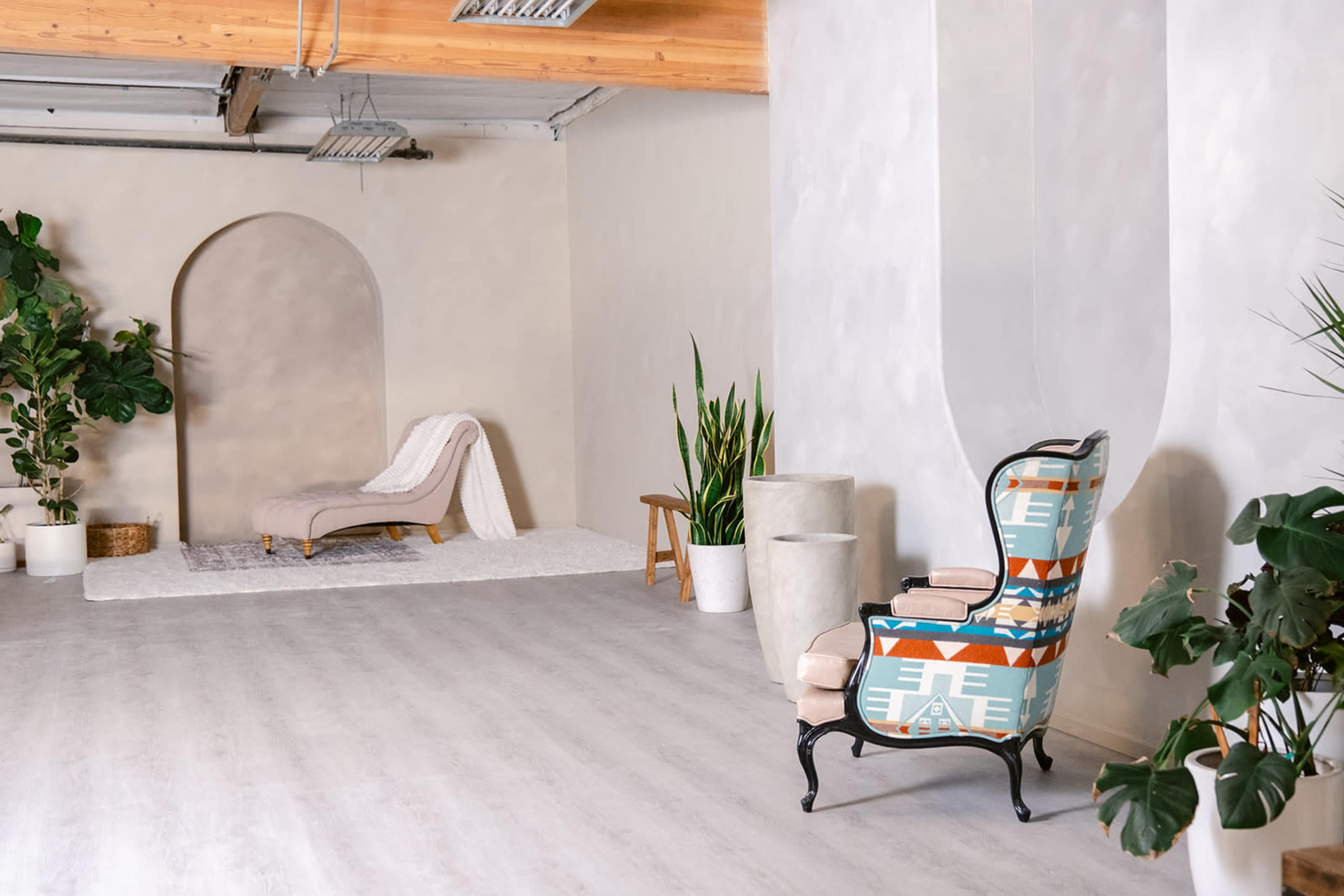 The image shows a modern interior space featuring a patterned armchair, a chaise lounge, potted plants, and neutral-colored walls.