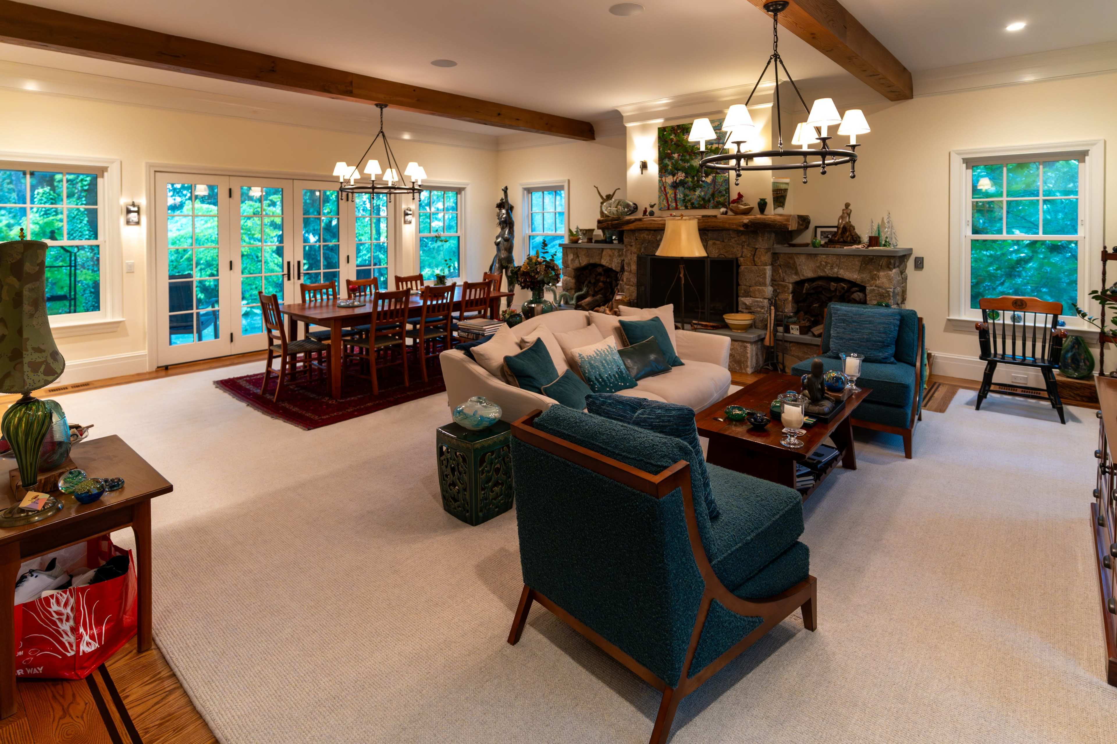 The spacious living area features a cozy seating arrangement facing a stone fireplace, with a dining table visible in the background surrounded by large windows.
