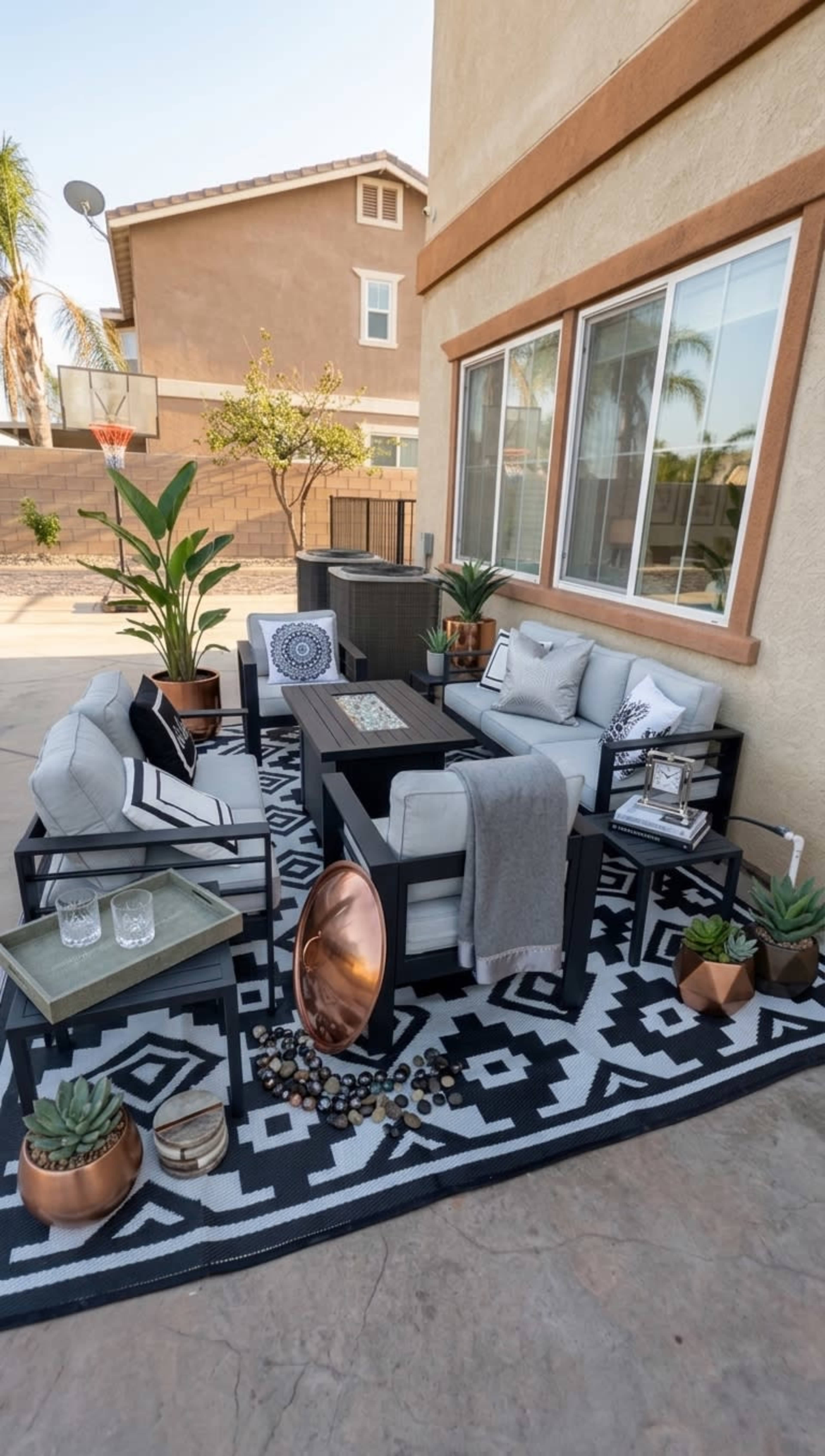 The image shows a stylish outdoor seating area featuring modern furniture, potted plants, and decorative elements arranged on a patterned rug.