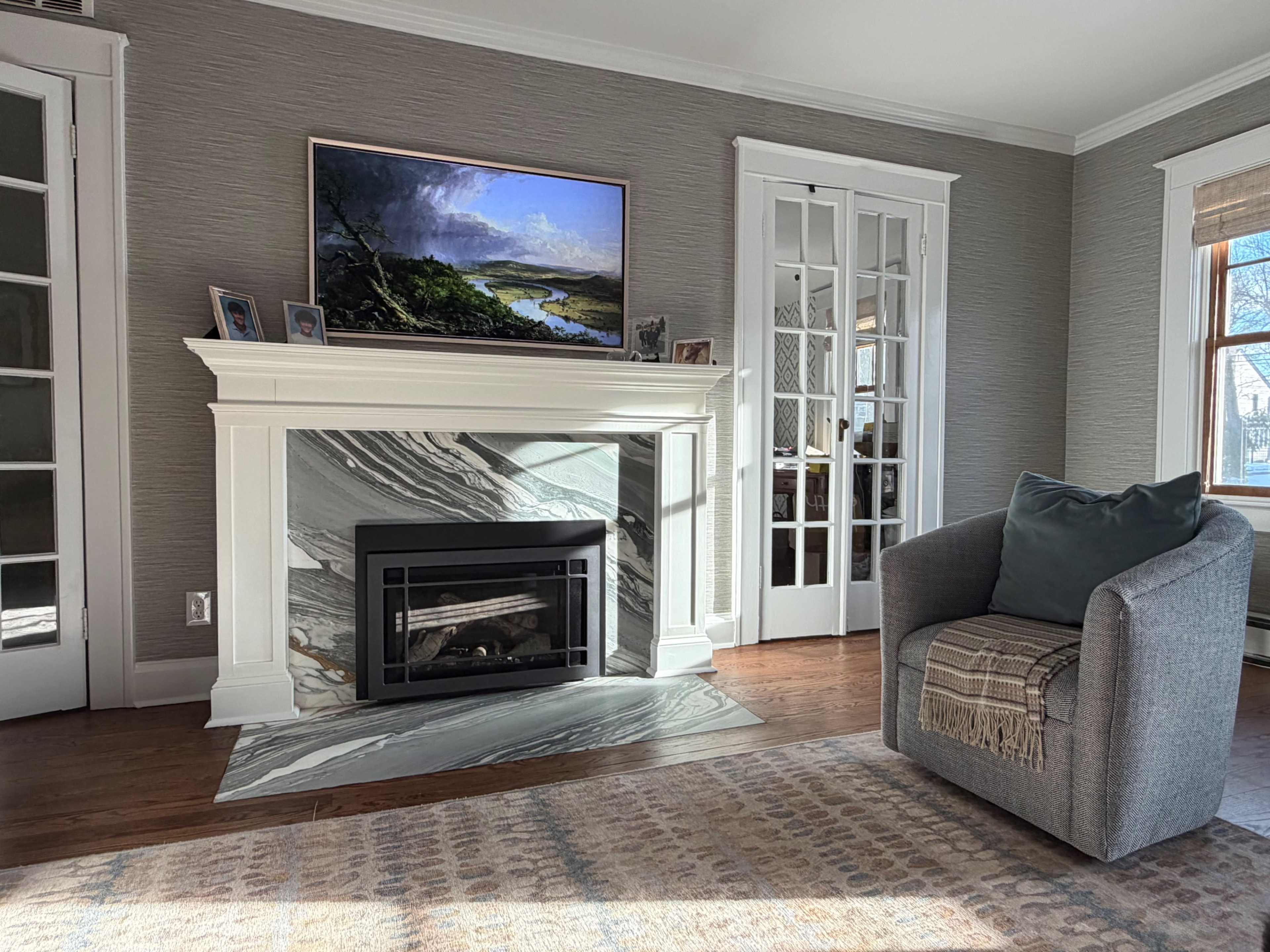 The image shows a living room with a fireplace, a mounted television displaying a landscape, a gray armchair, and light-colored walls.