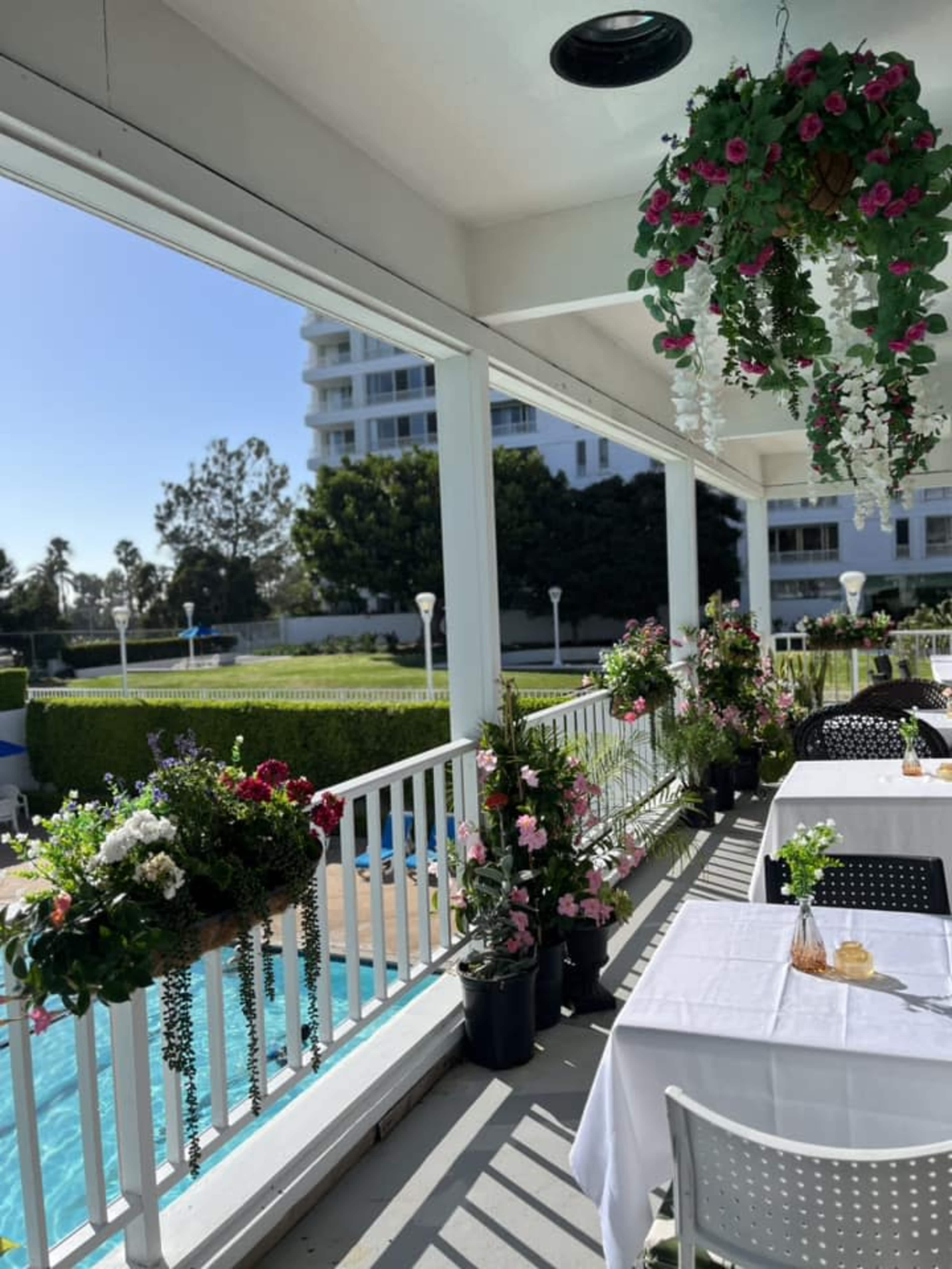 A dining area with white tablecloths is set along a patio overlooking a swimming pool, adorned with hanging floral arrangements and greenery.