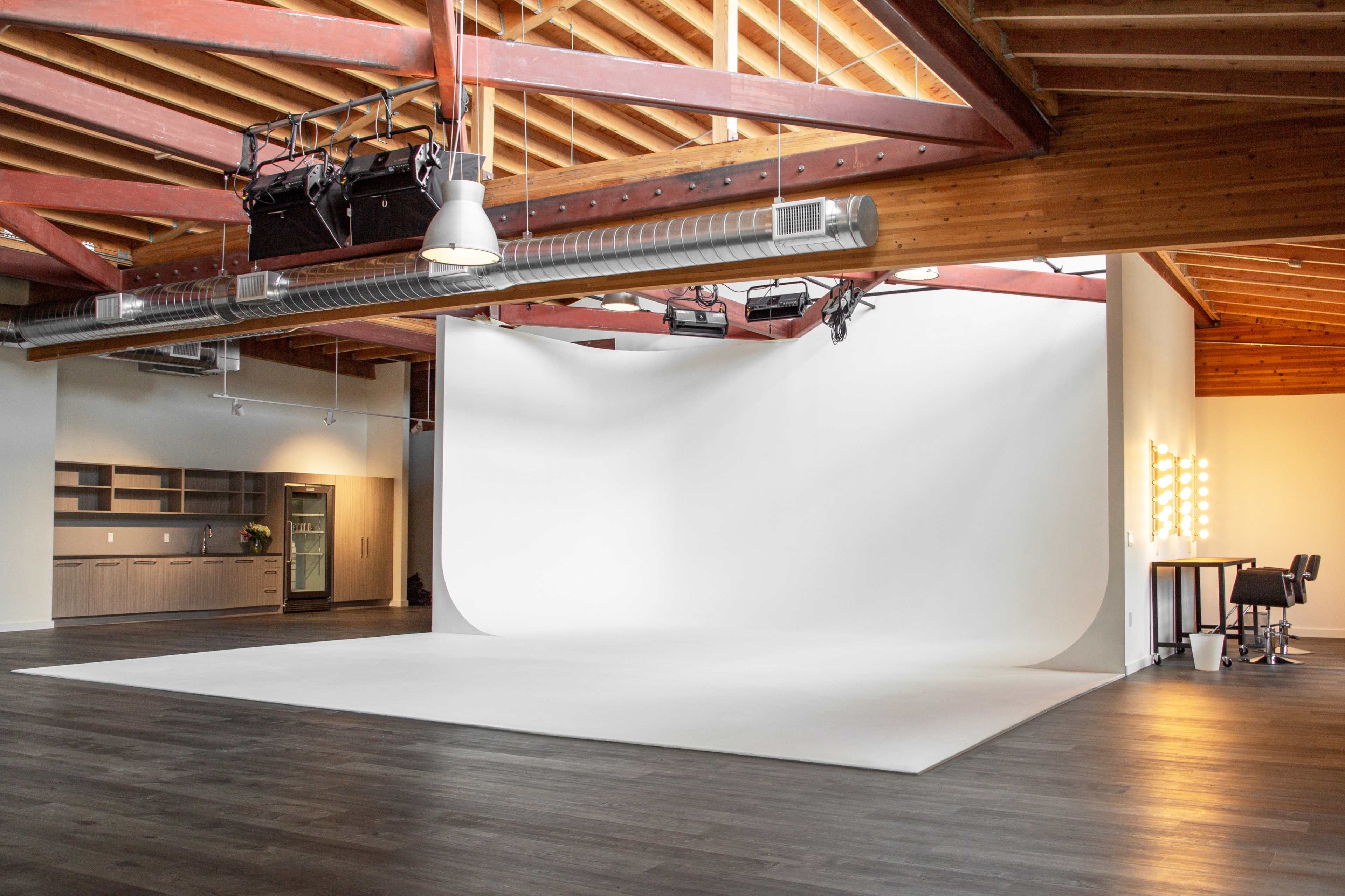 A spacious photography studio with a seamless white backdrop, wooden beams, and a large mirrored vanity area.