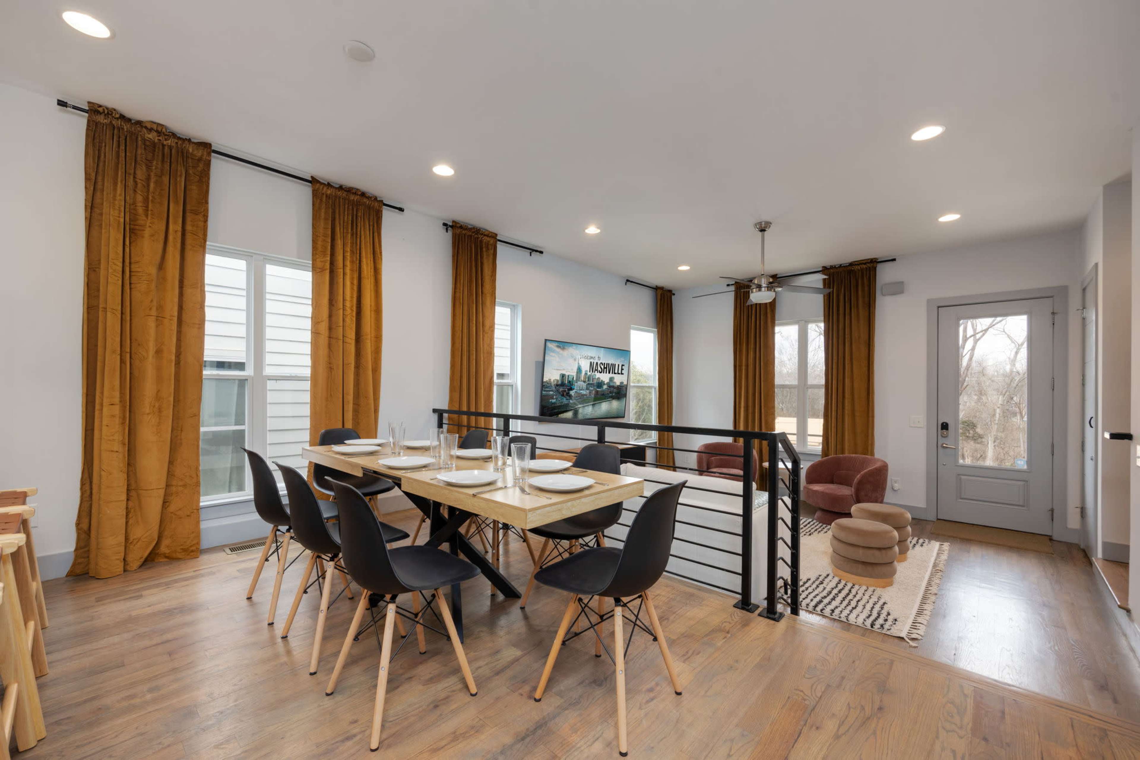 A modern dining area features a rectangular table set for six, surrounded by black chairs, with large windows dressed in golden curtains and a brown armchair in the corner.