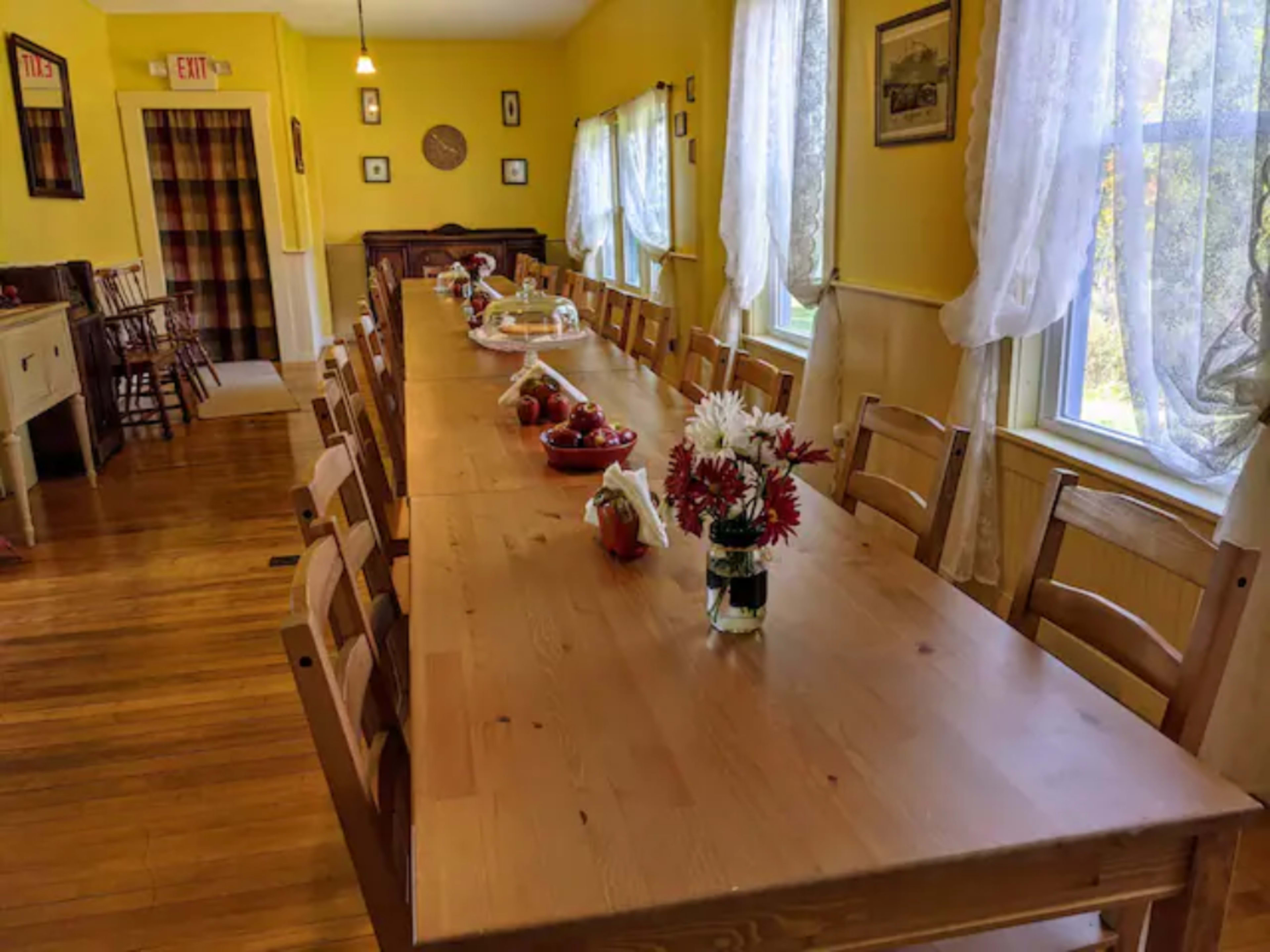 A long wooden dining table is set with fruit and flowers in a brightly lit room with yellow walls and large windows.