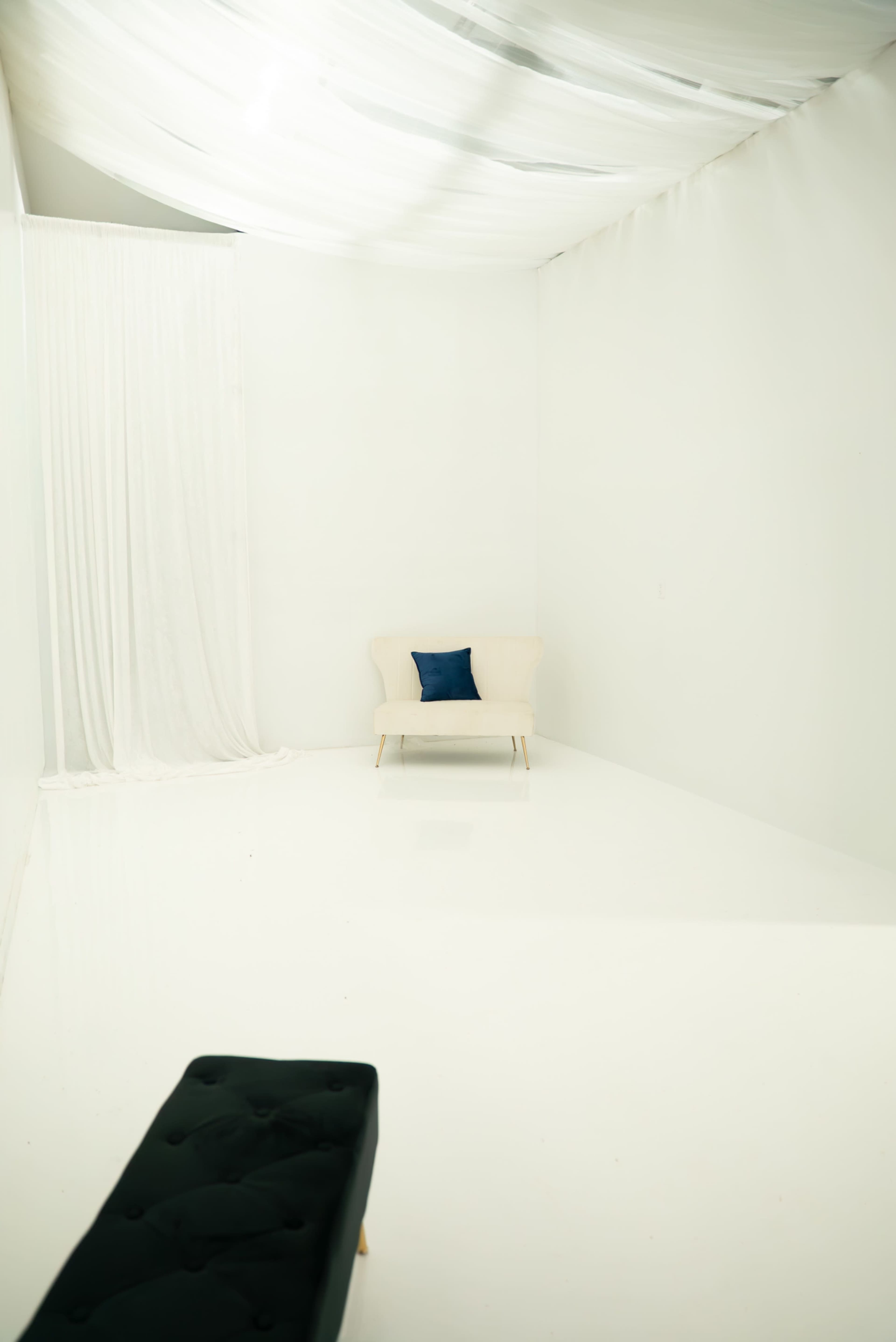 The image shows a minimalistic, white room with a single beige chair and a dark blue cushion, alongside sheer curtains draping from one wall.