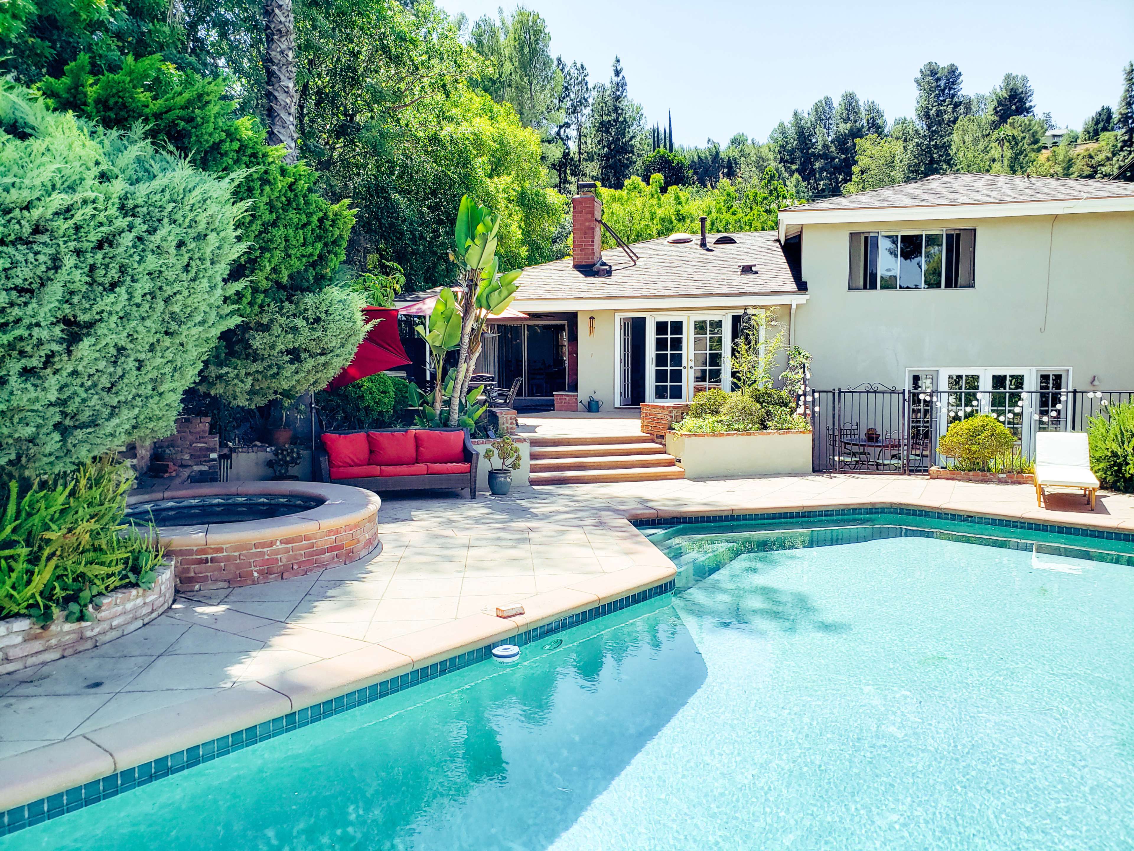 The image shows a swimming pool in the foreground with a residential patio and garden area in the background, featuring a house surrounded by greenery.