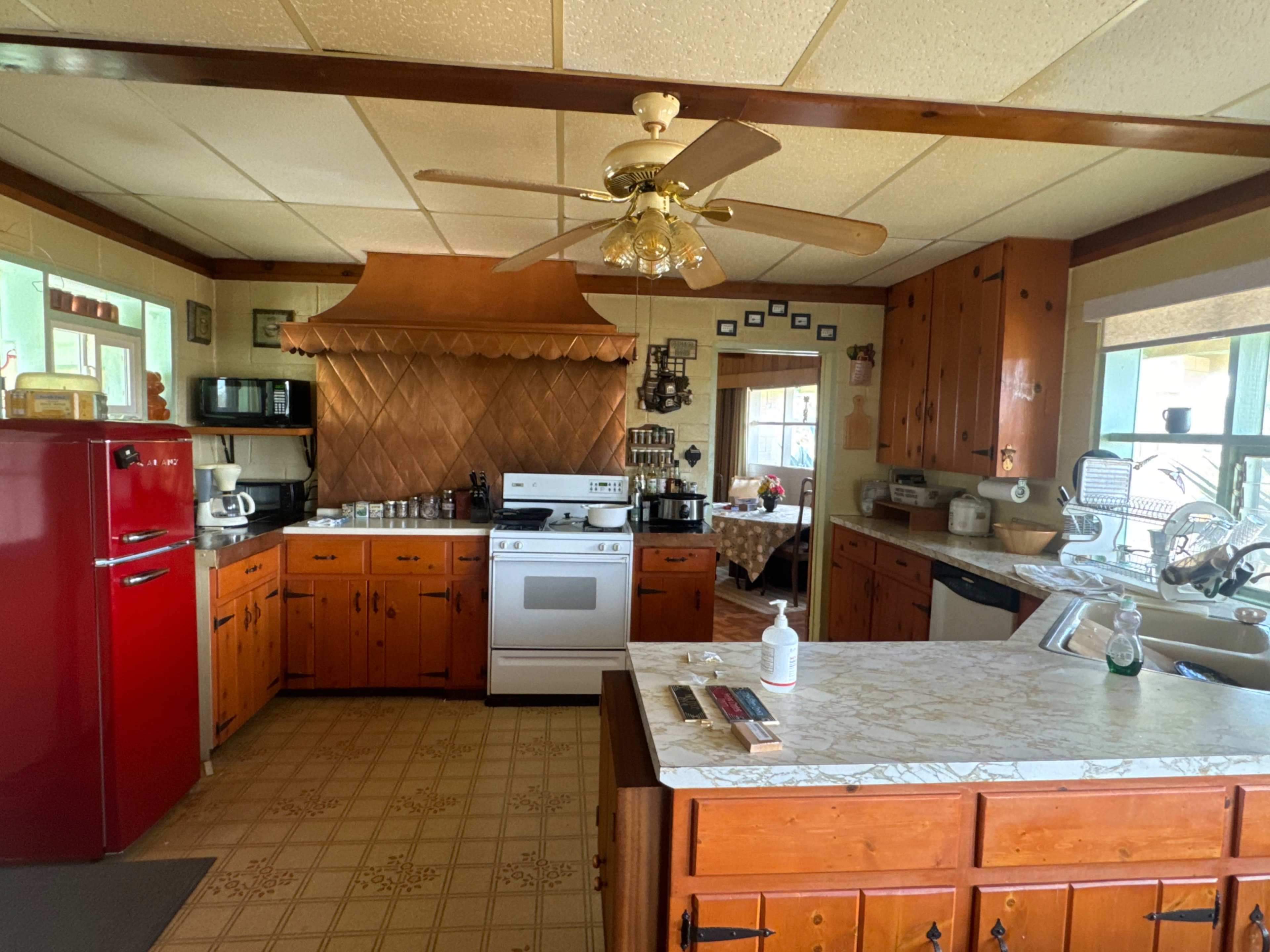 The kitchen features wooden cabinets, a red retro refrigerator, and various appliances arranged around a central island with a marble countertop.