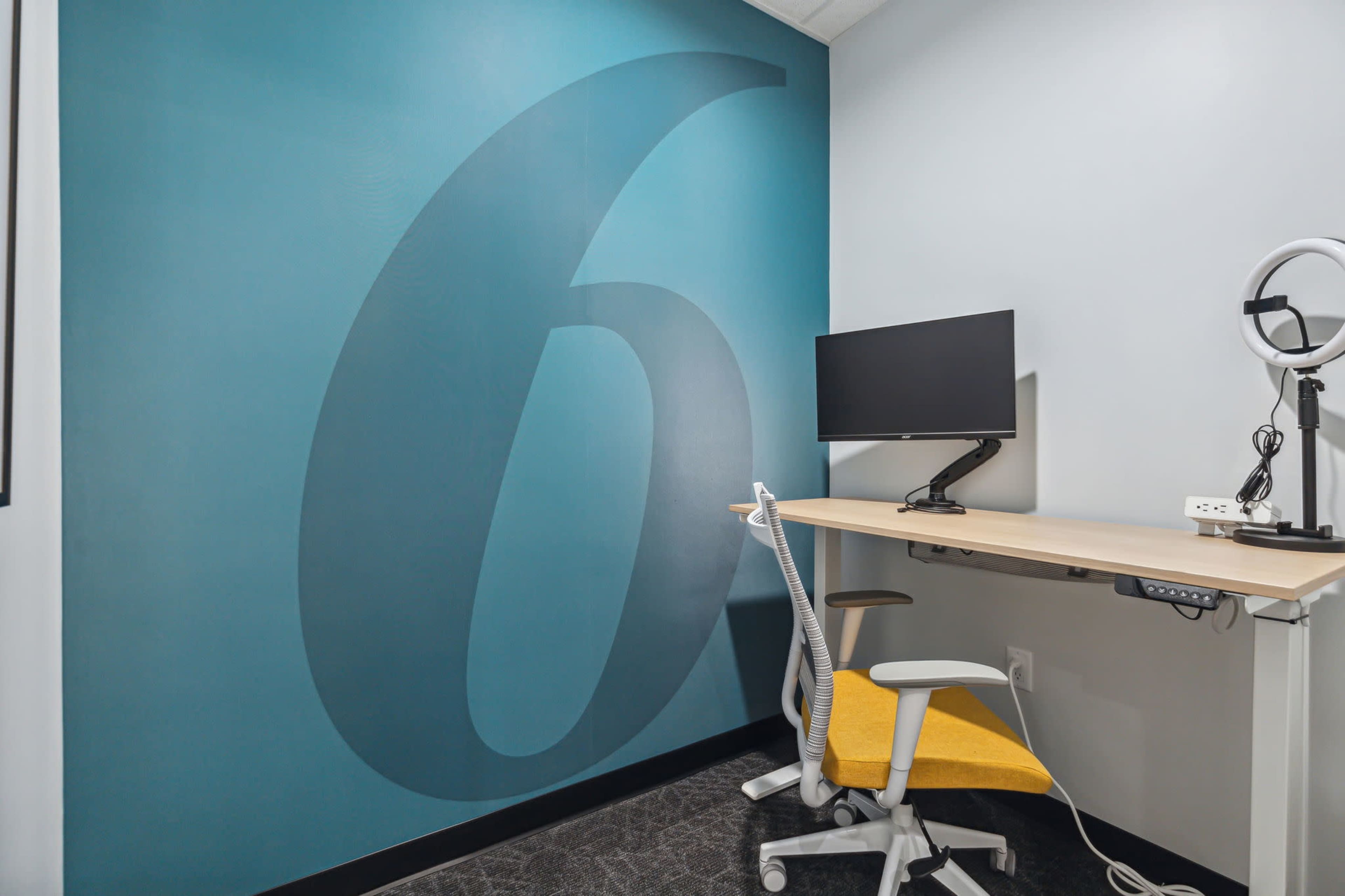 The image shows a small workspace featuring a desk with a monitor, a lamp, and a chair, against a wall painted with the number six.