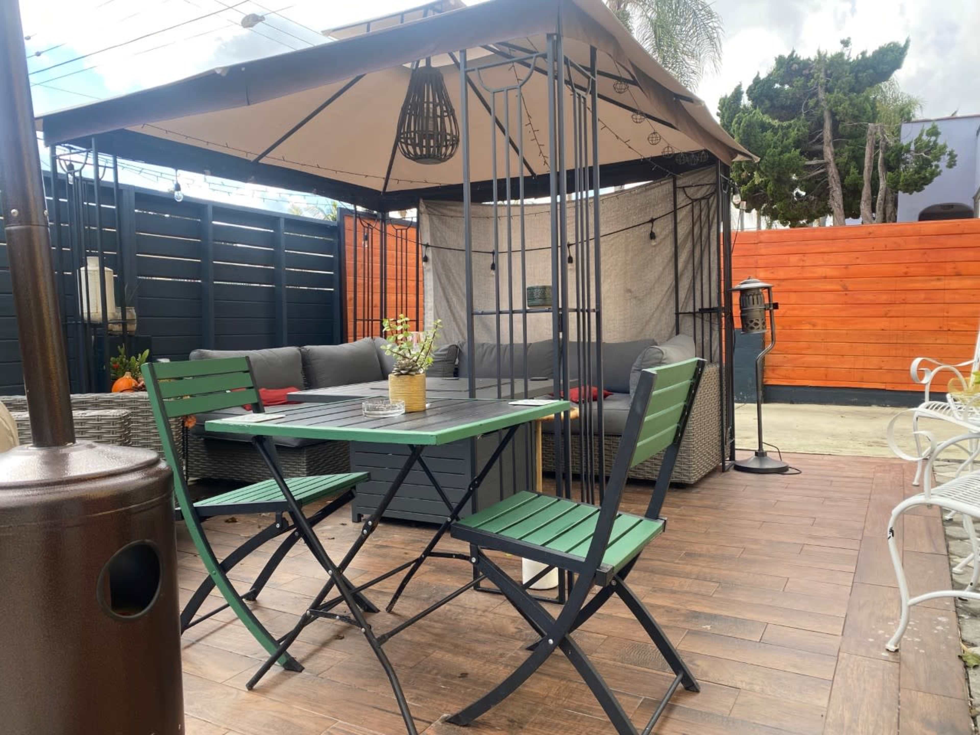 A patio area featuring a gazebo, a table with green chairs, and a seating arrangement with gray cushions surrounded by wooden fencing.