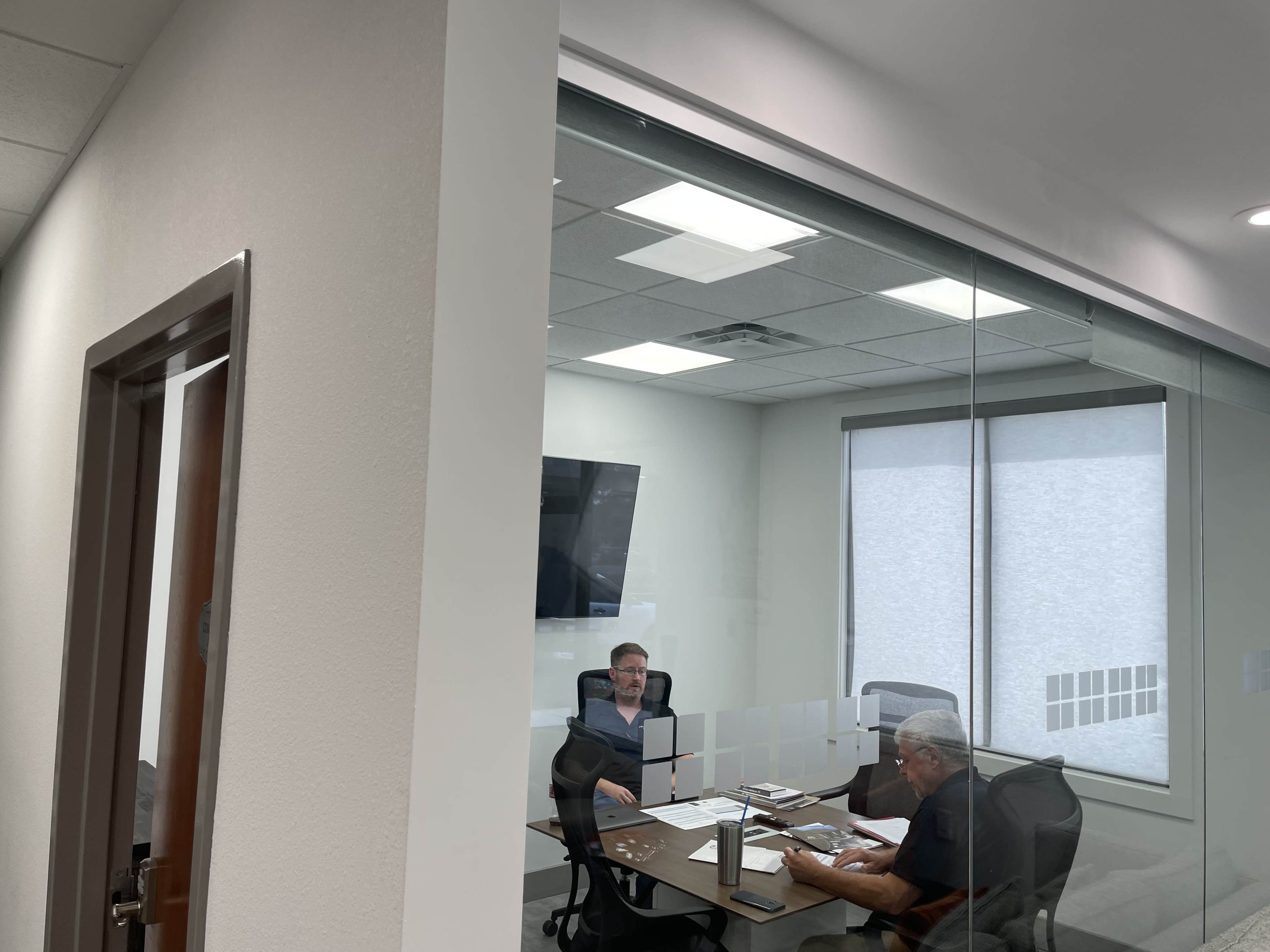 Two men are seated at a table in a glass-walled meeting room, with a television on the wall and documents spread out in front of them.