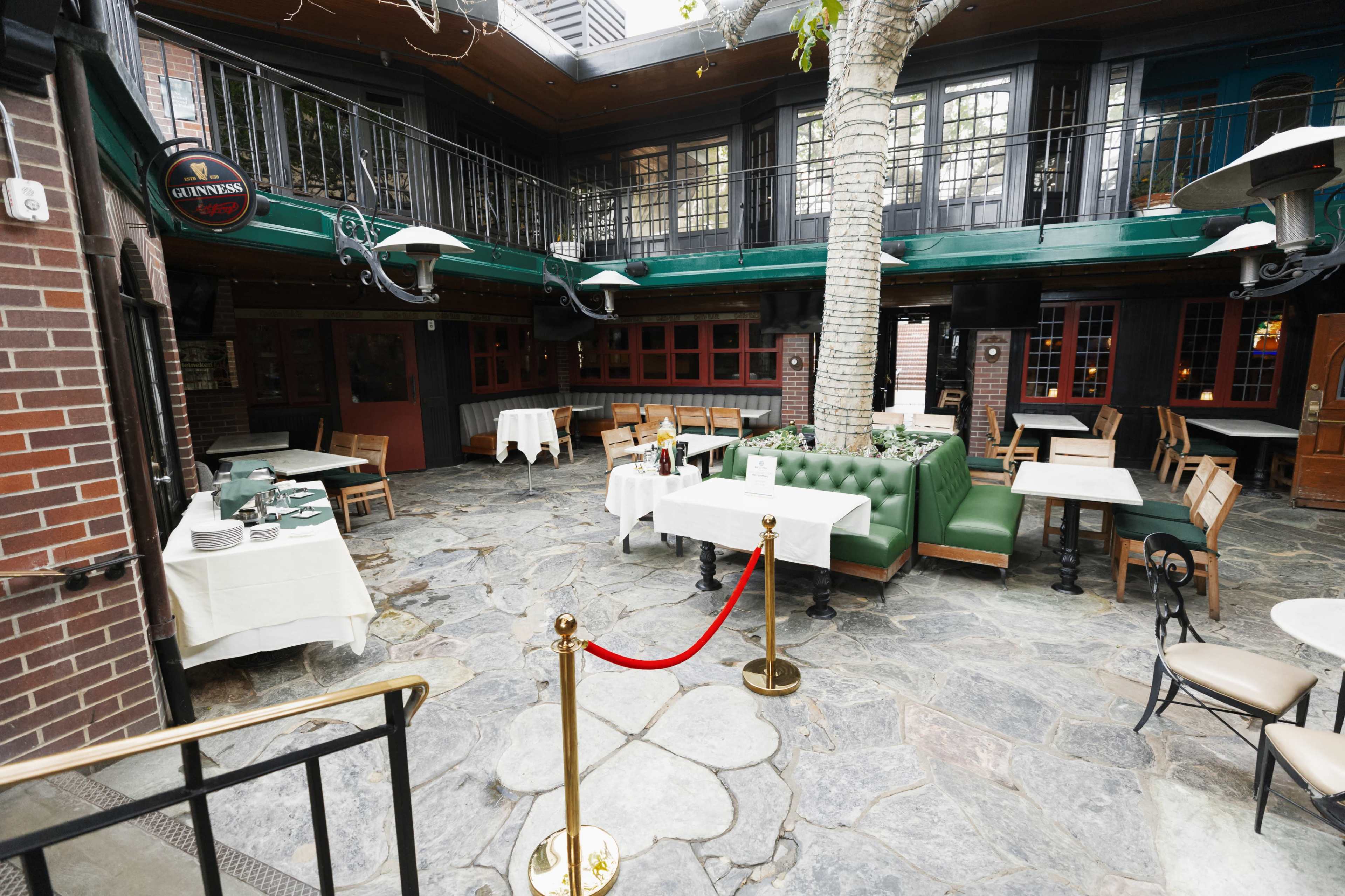 The image shows a spacious restaurant with a stone floor, green upholstered seating, tables covered with white tablecloths, and a tree situated in the center.