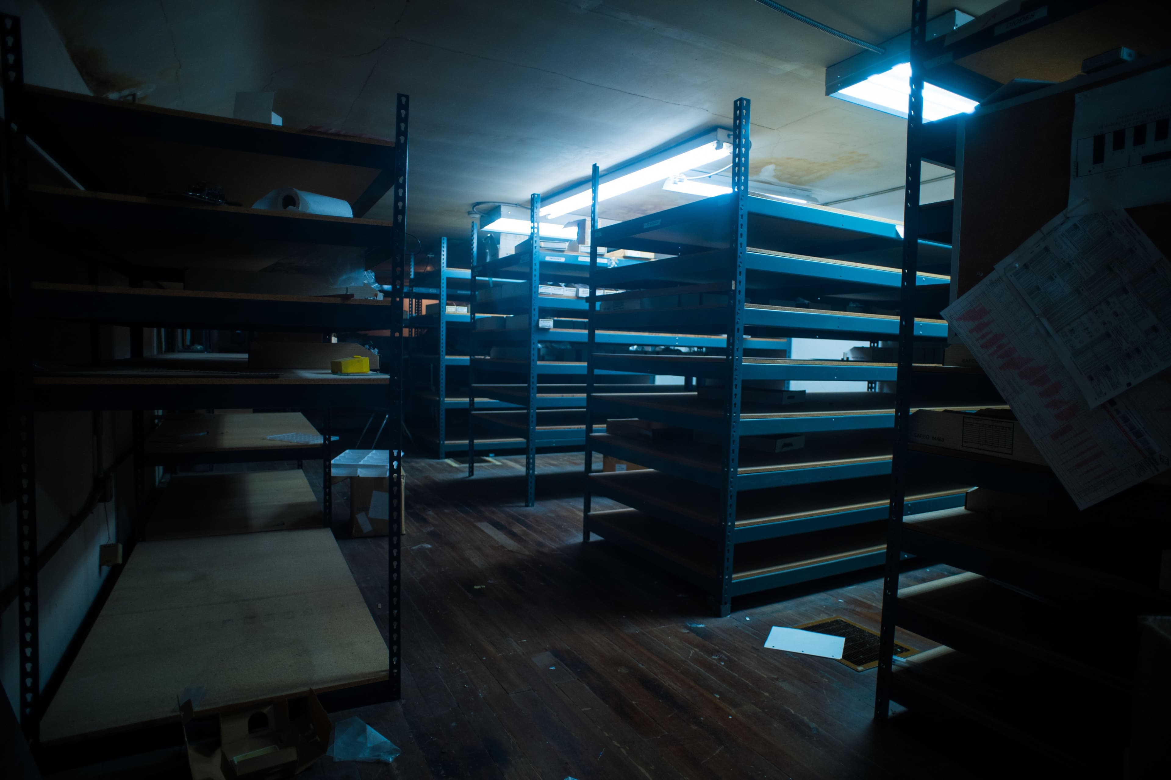 A dimly lit room with empty metal shelves organized in rows, revealing a wooden floor scattered with debris.