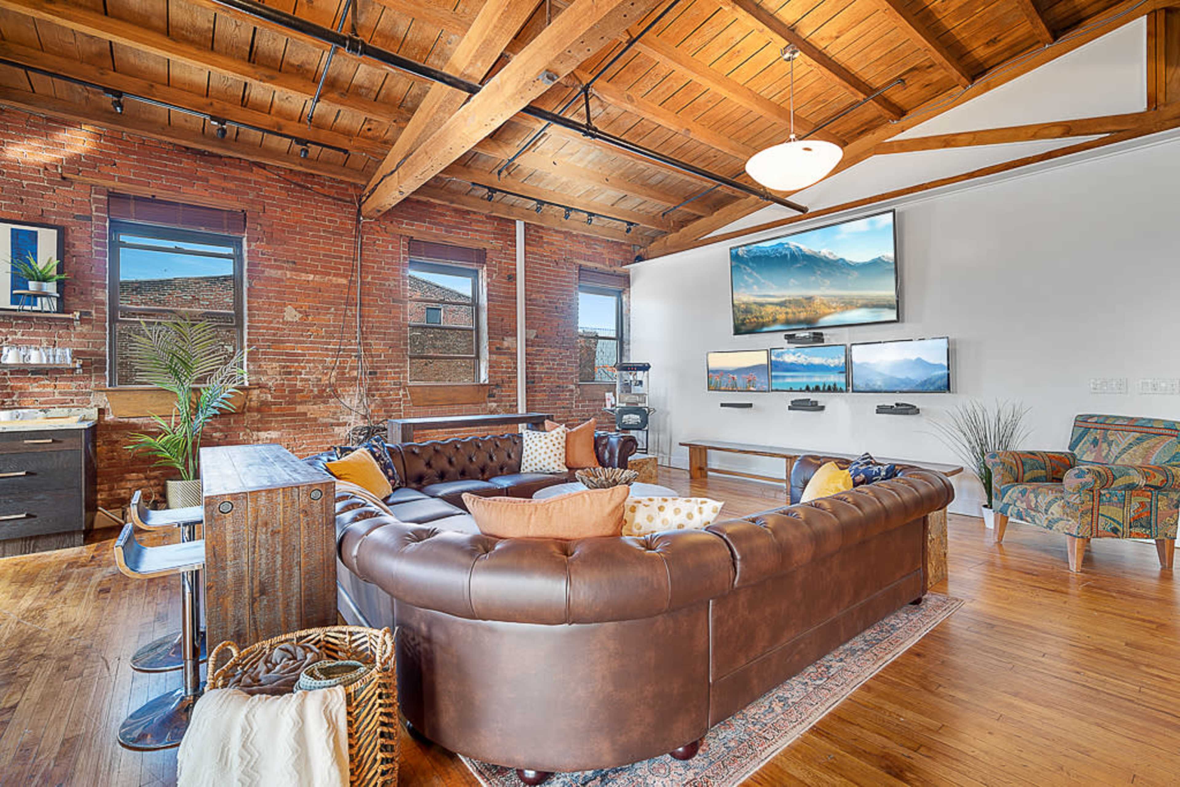 A modern living room with exposed brick walls, wooden beams, and a large leather sectional sofa arranged around a coffee table, complemented by multiple mounted TVs on the wall.