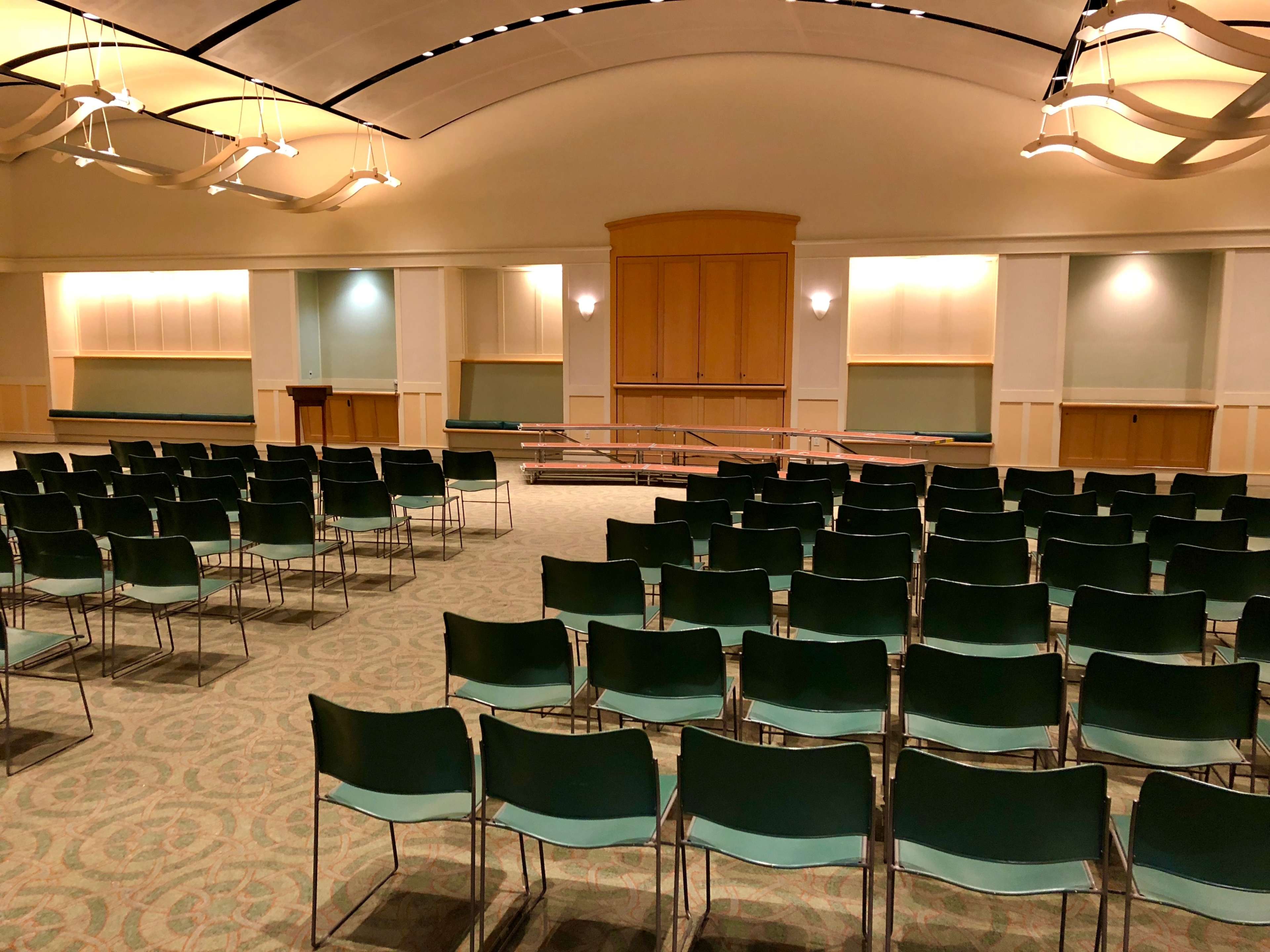 A spacious auditorium is set up with rows of green chairs facing a wooden stage at the front.