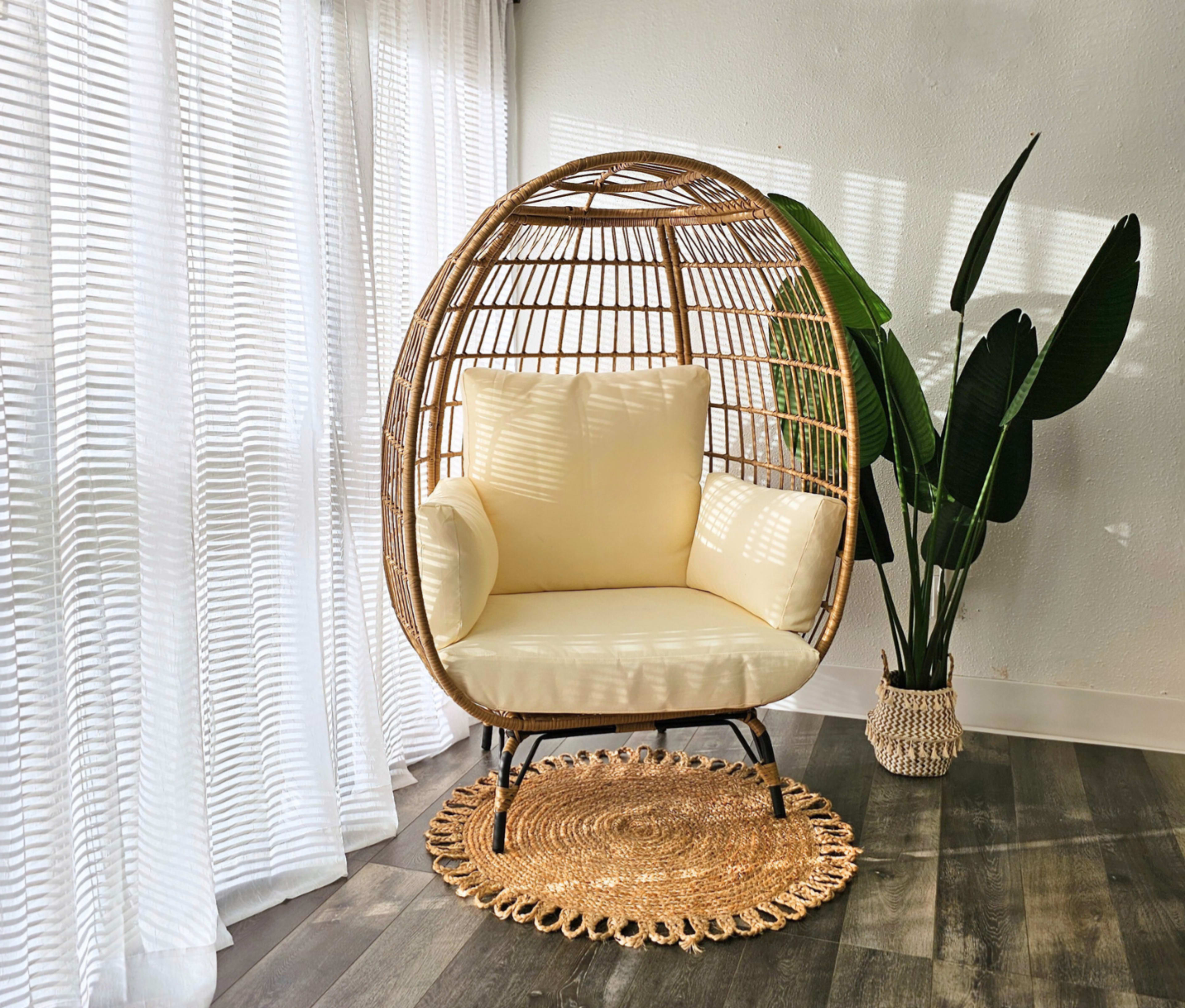 A round, woven chair with cream cushions sits next to a leafy plant on a textured rug in a room with sheer white curtains.