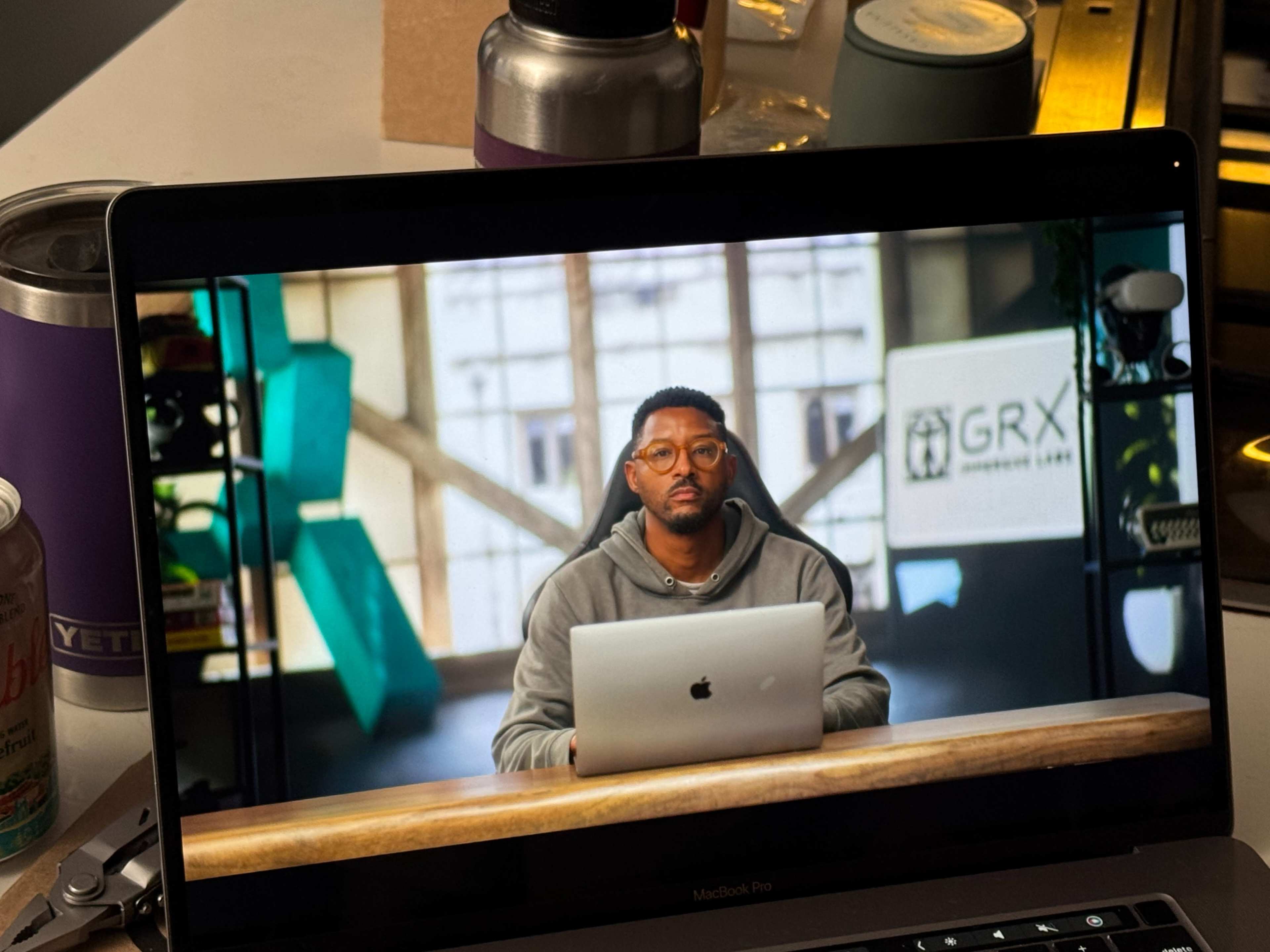 A person with glasses sits at a desk in front of a laptop, with a large screen displaying the "GRX" logo in a well-lit room.