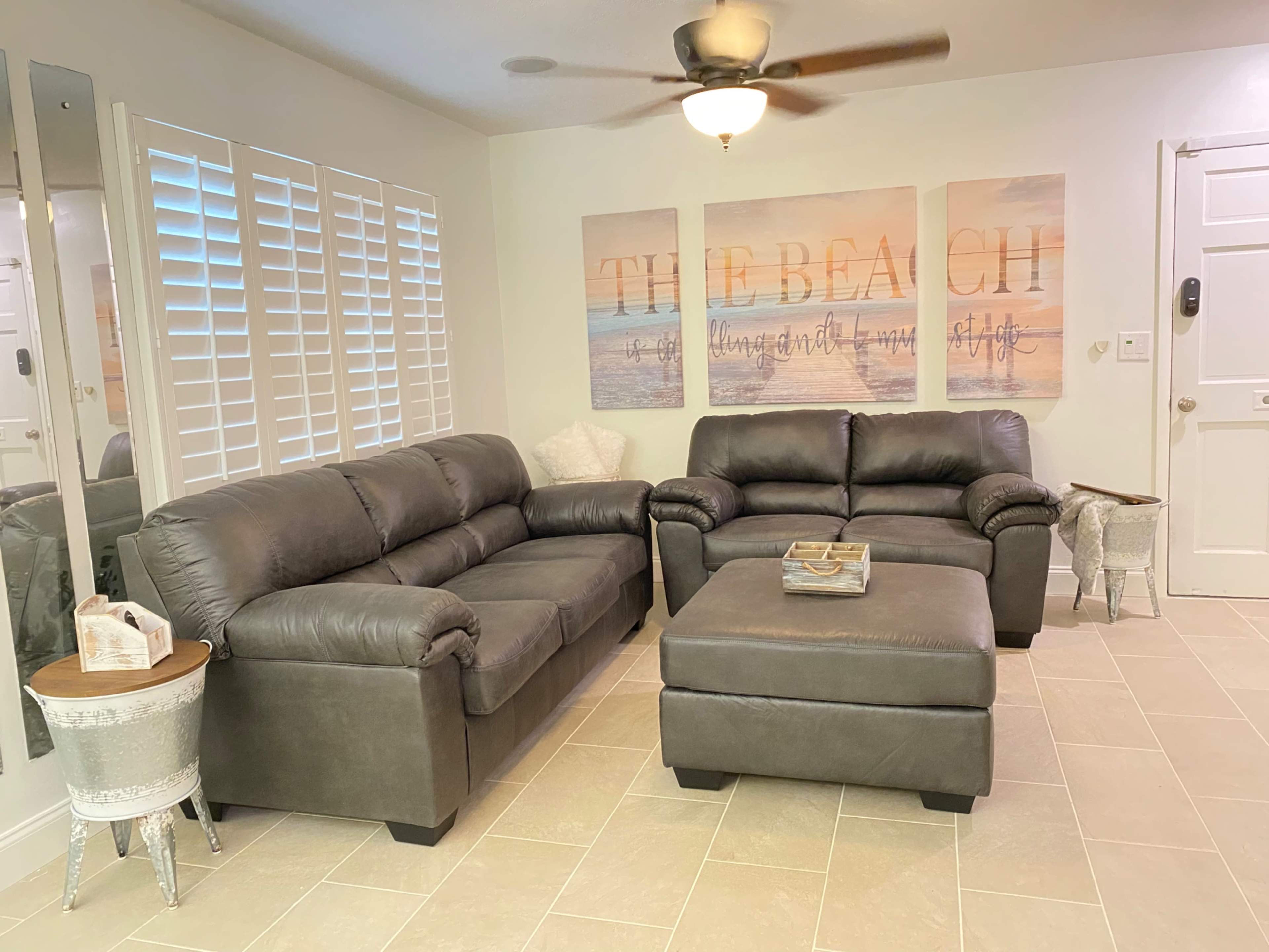 The living room features a gray leather sofa and loveseat arranged around an ottoman, with beach-themed artwork on the wall and a door leading outside.