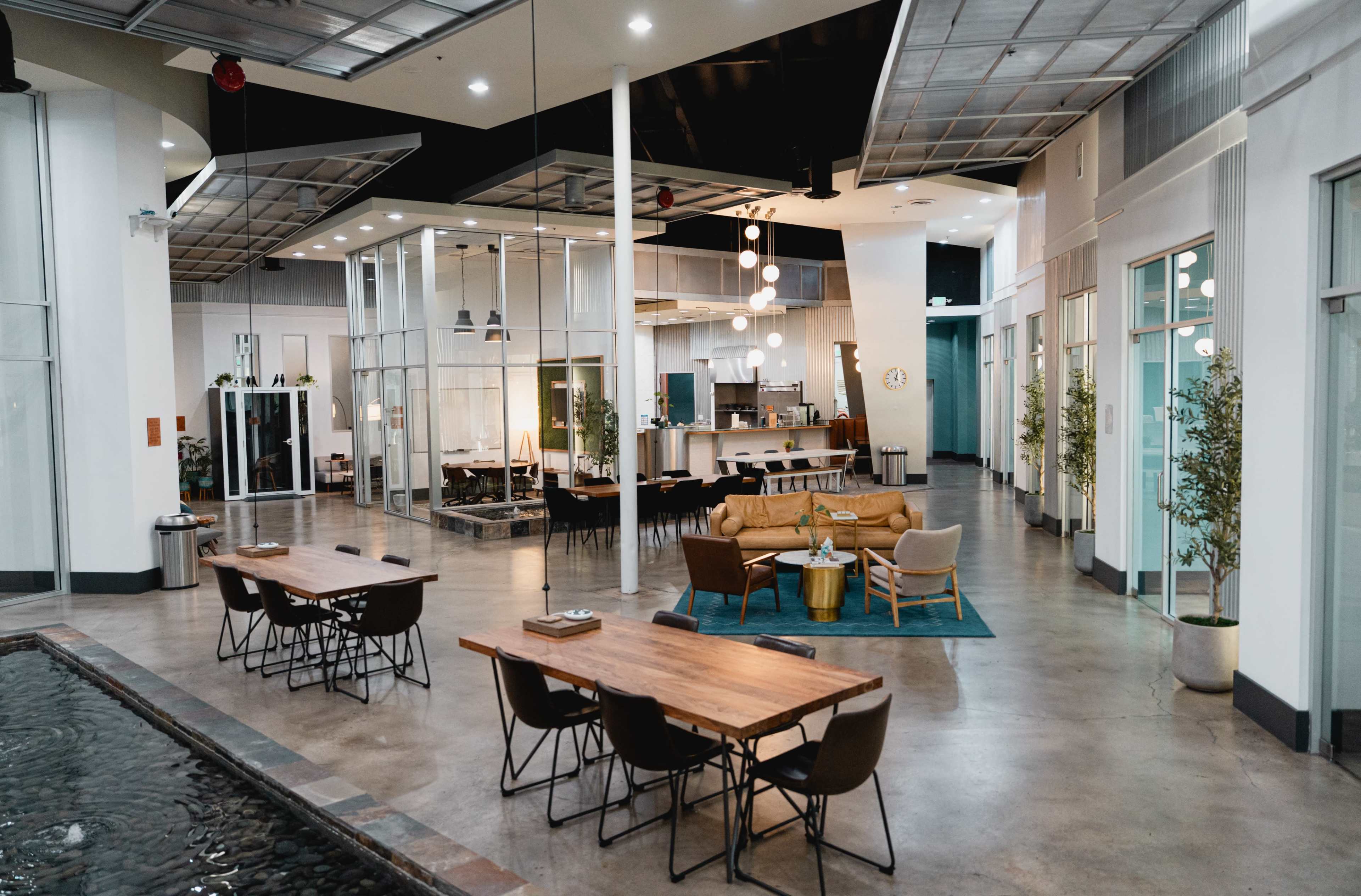 The image shows a modern, open-concept communal space featuring various seating arrangements, a kitchen area, and large windows letting in natural light.