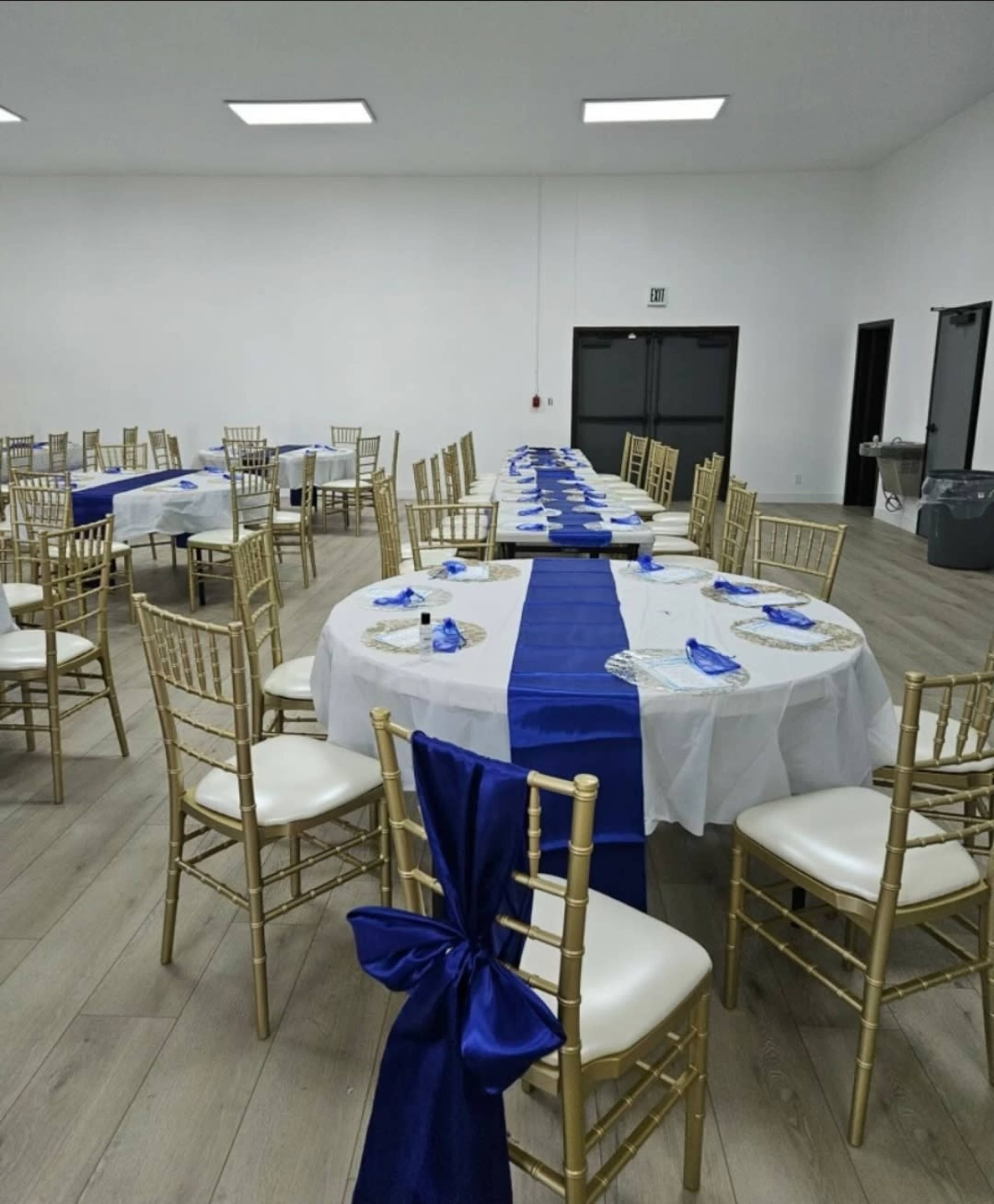 A banquet hall is set up with tables covered in white tablecloths and royal blue runners, surrounded by gold Chiavari chairs.