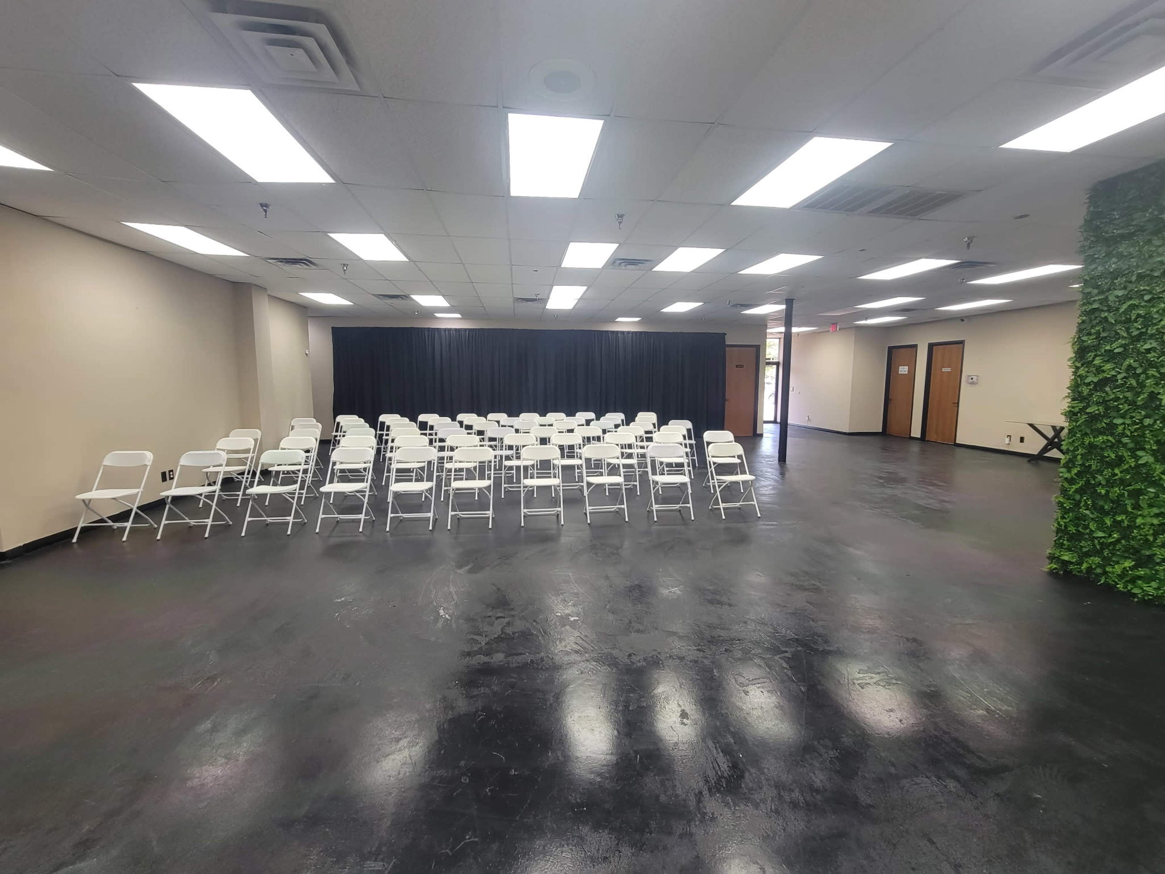 The image shows an empty room with rows of white folding chairs facing a black curtain, surrounded by plain walls and a green plant wall on one side.