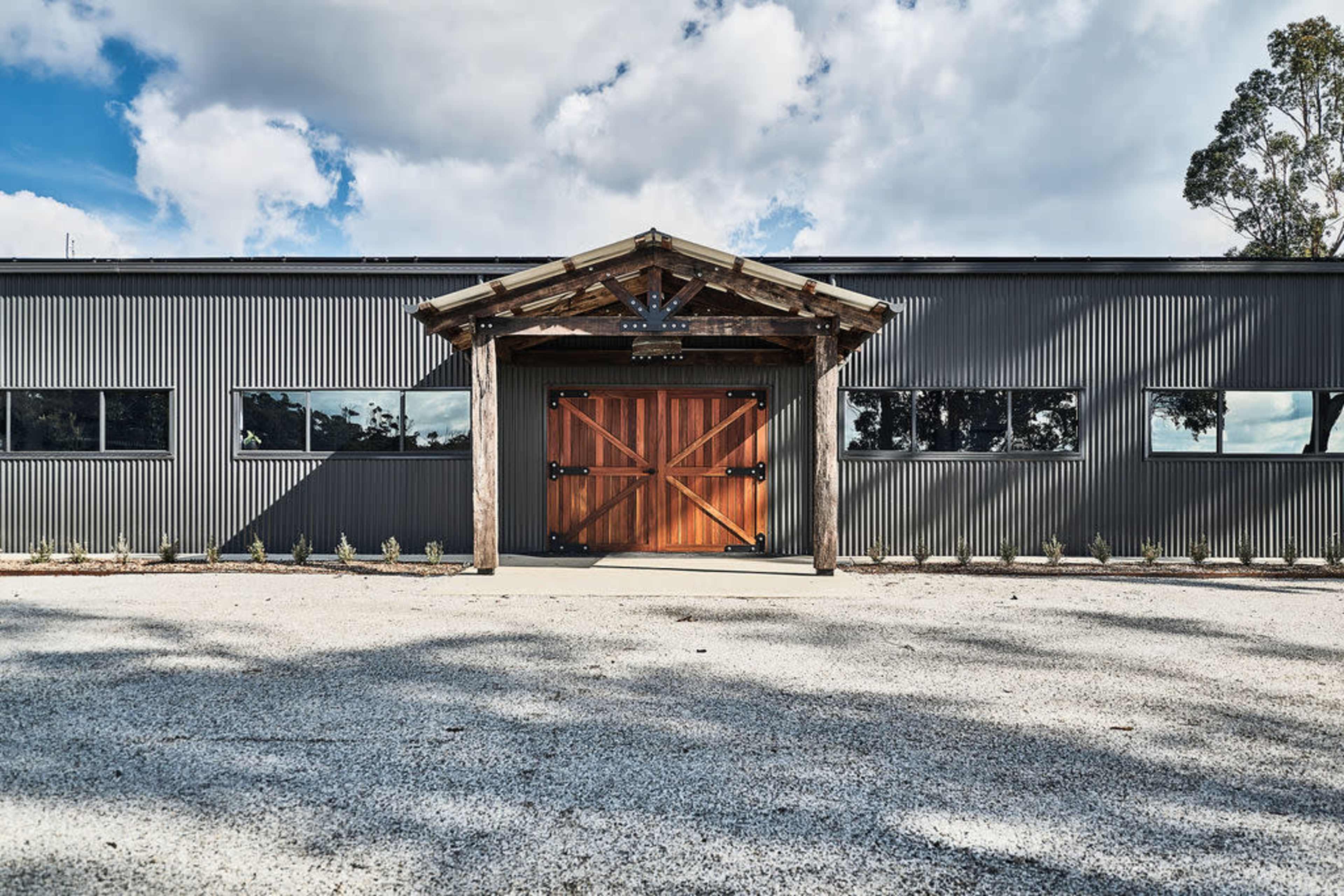 A modern building features a large wooden entrance flanked by multiple windows, set against a gravel parking area.