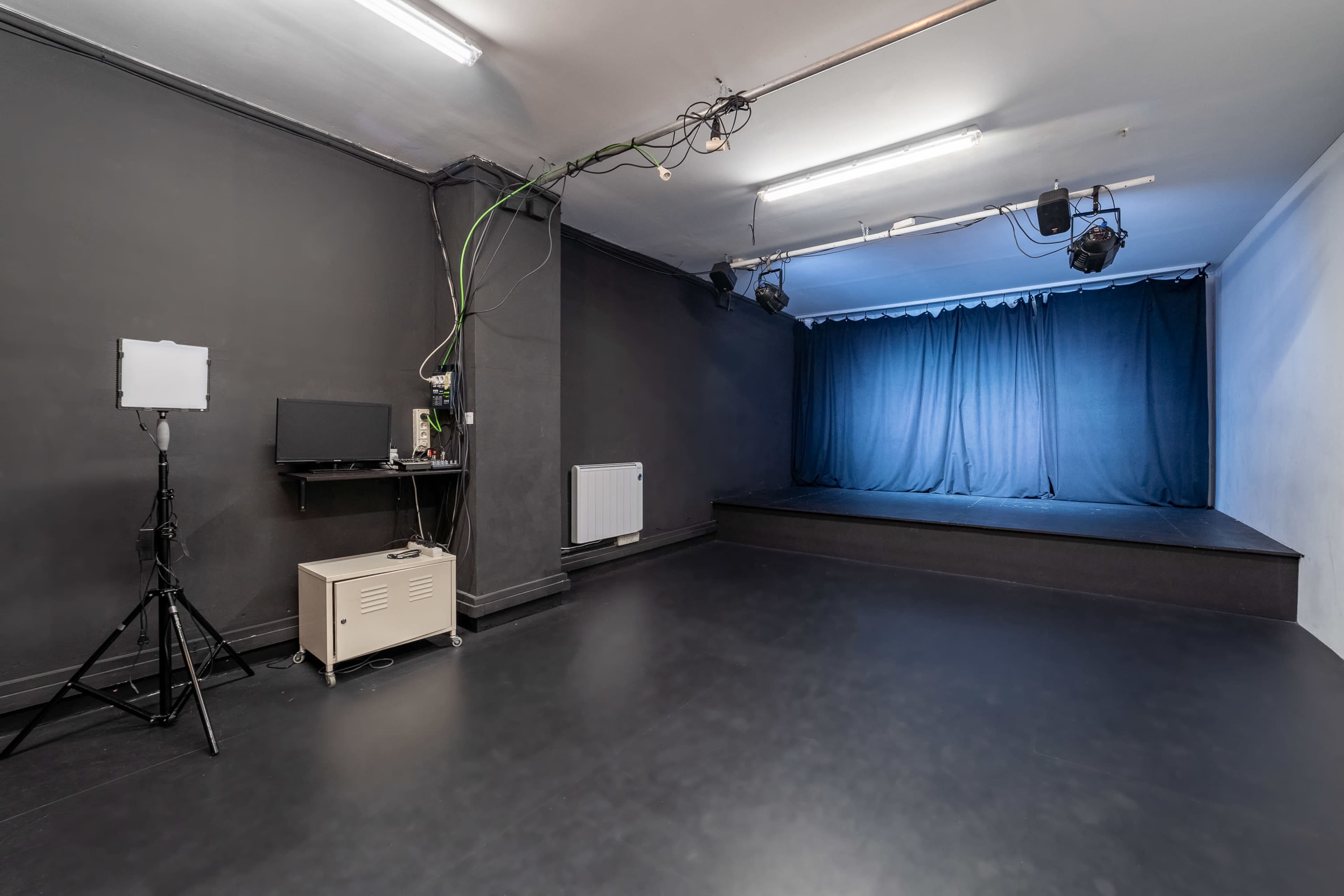 A sparse performance space with a blue curtain, simple lighting, a small stage, and minimal equipment along one wall.