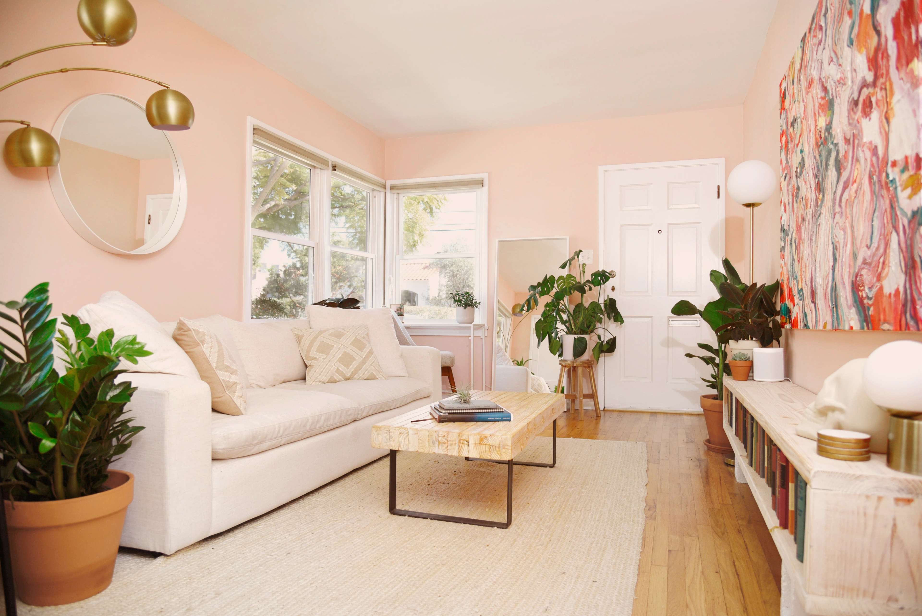 A living room with a light pink wall, a white sofa, a wooden coffee table, indoor plants, and artwork on the wall.