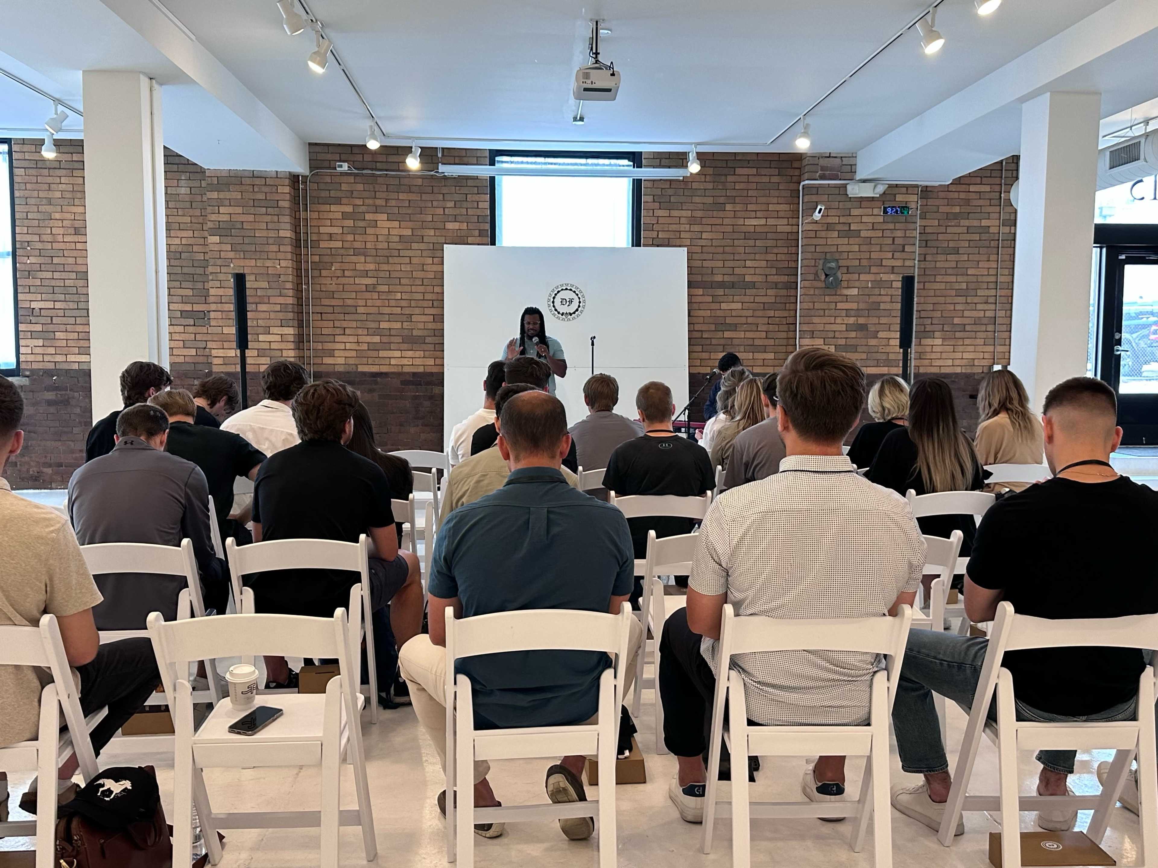 A speaker addresses an audience seated in white chairs in a brick-walled event space.