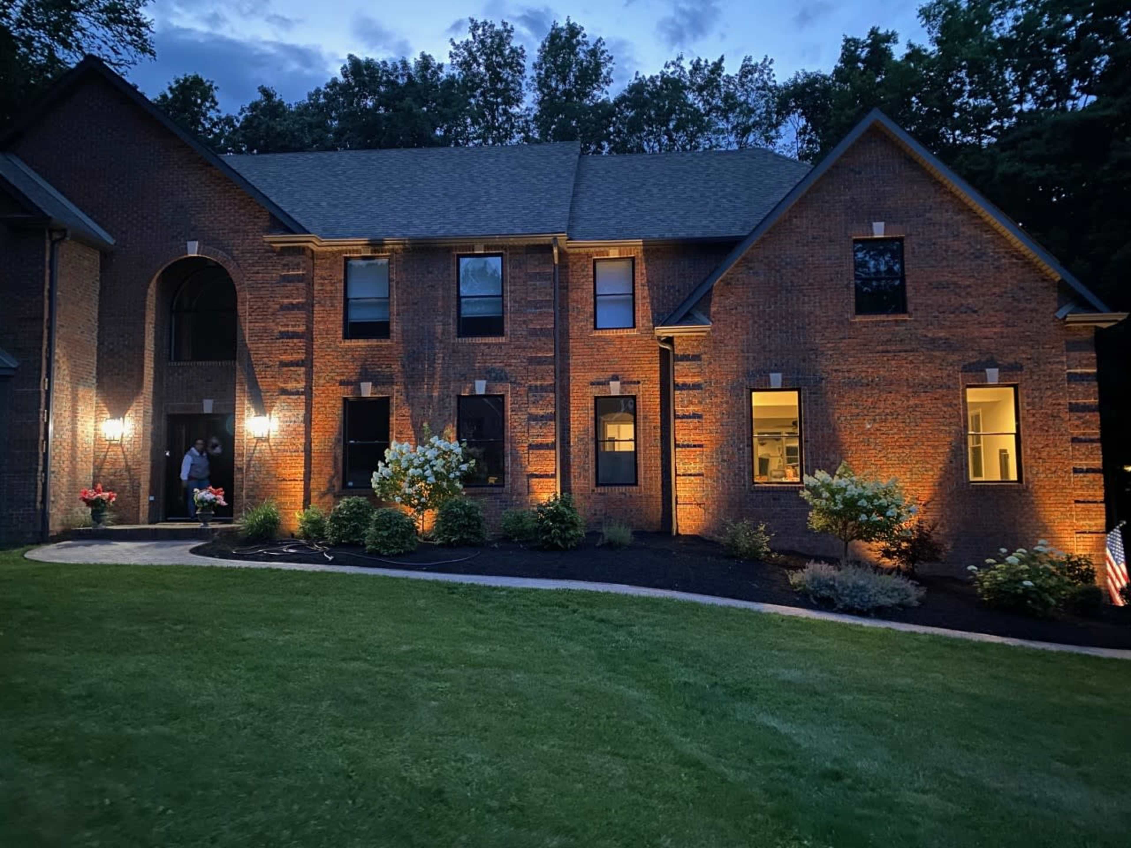A two-story brick house is illuminated at dusk, surrounded by a well-maintained lawn and colorful flower beds.
