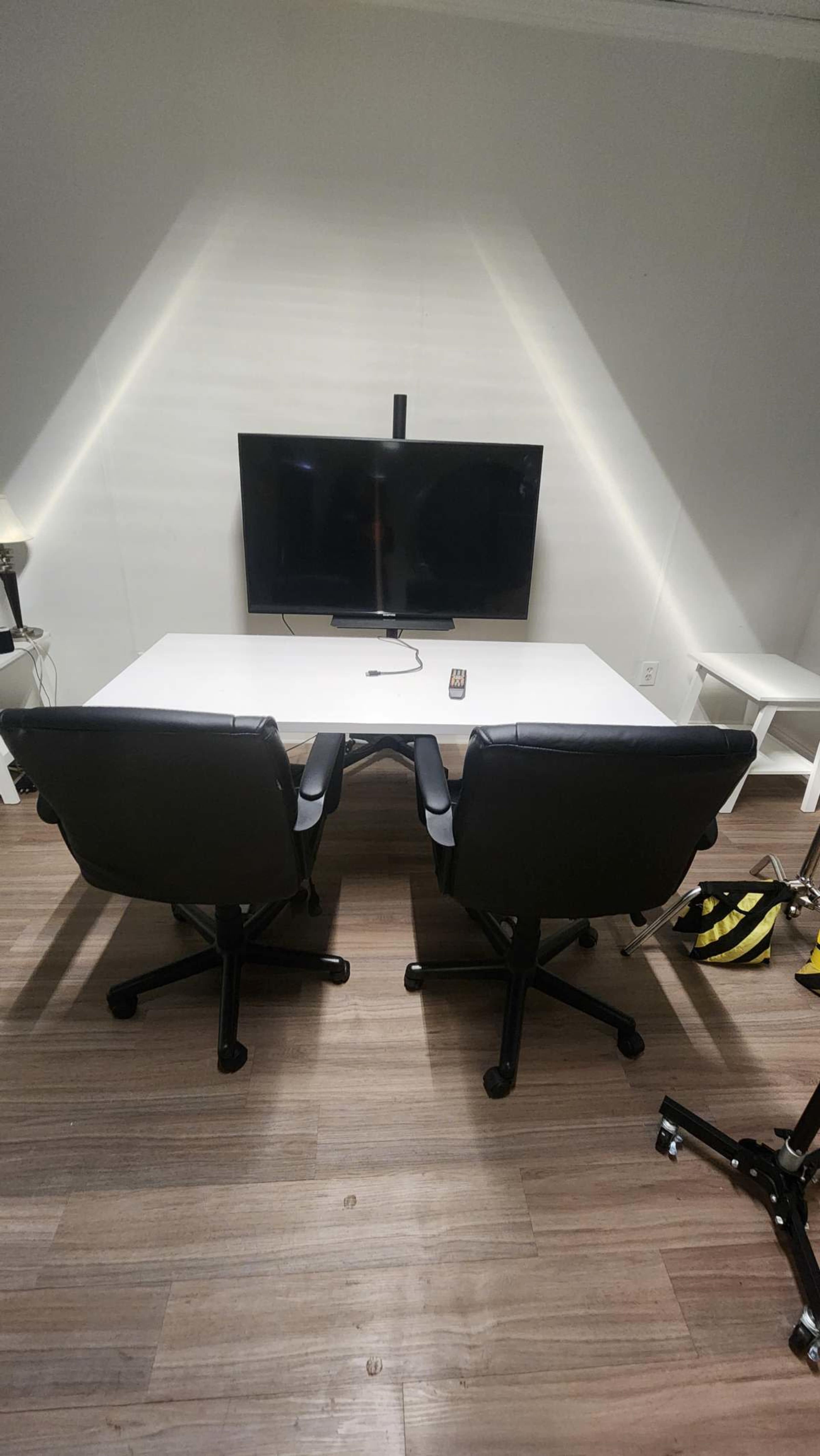 The image shows a conference room with a white table, two black rolling chairs, and a wall-mounted television.