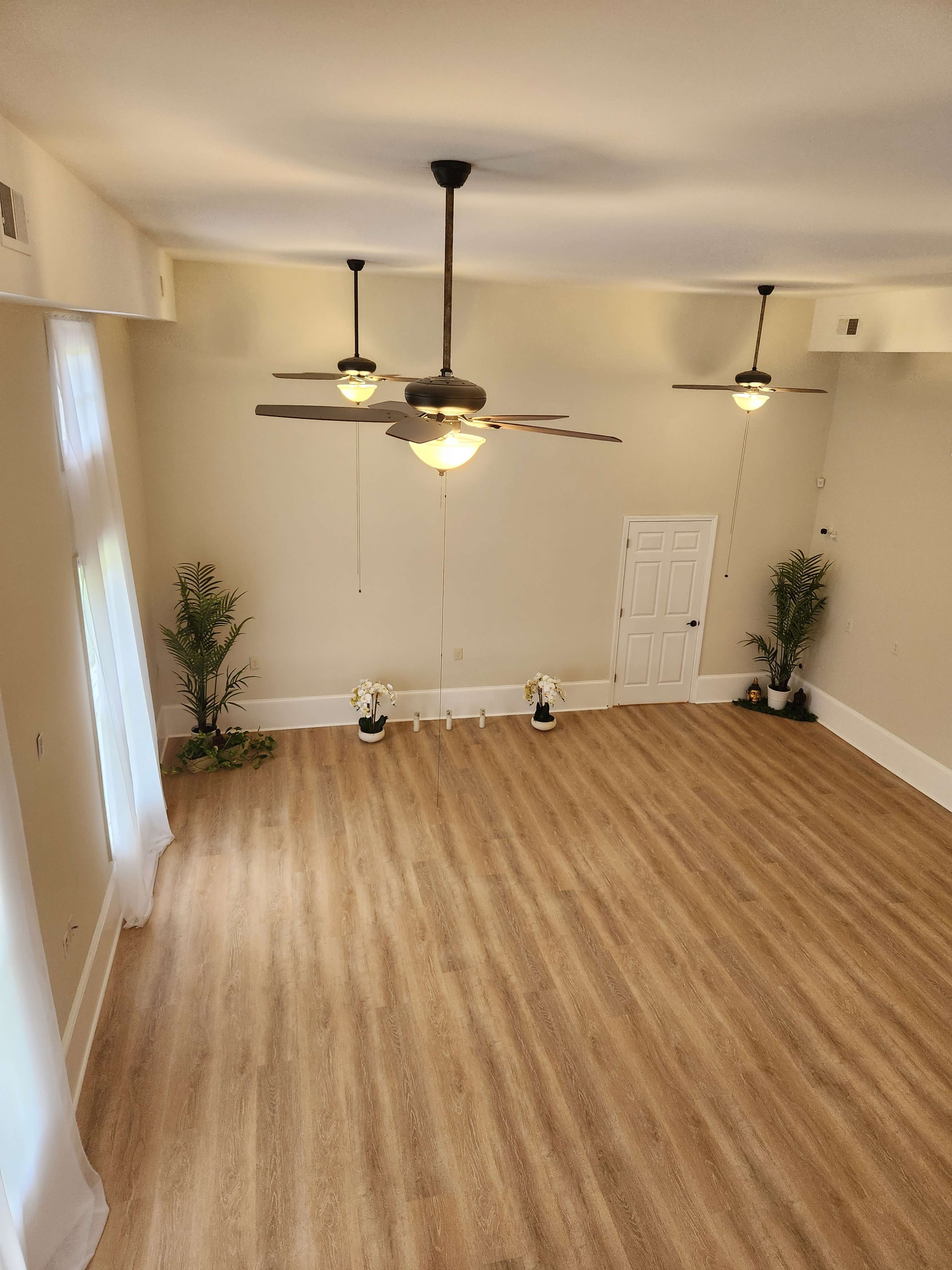 A spacious, empty room with light wooden flooring, two ceiling fans, and potted plants in the corners.
