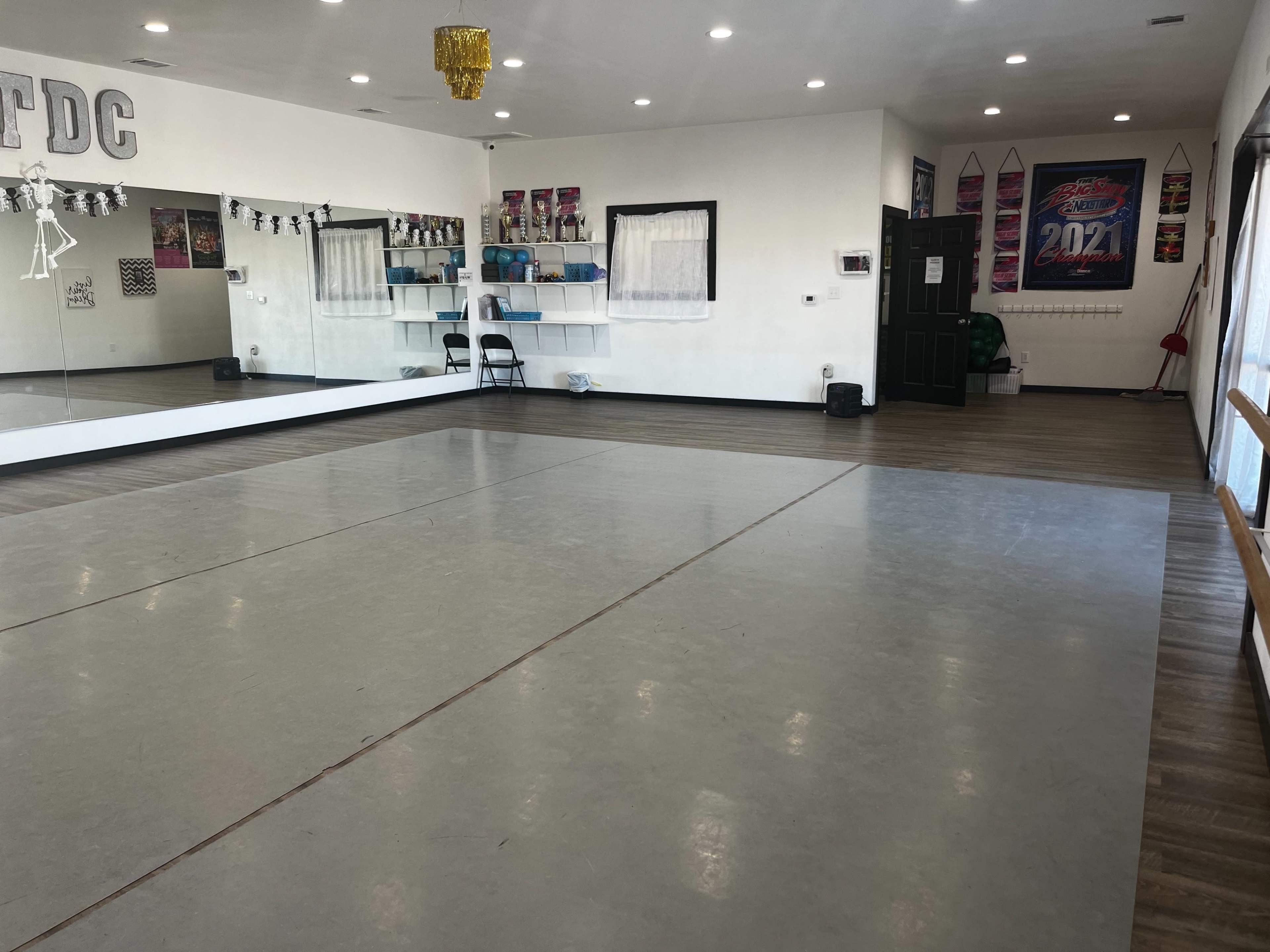 The image shows a spacious dance studio with a mirrored wall, wooden floors, and shelves lined with equipment.