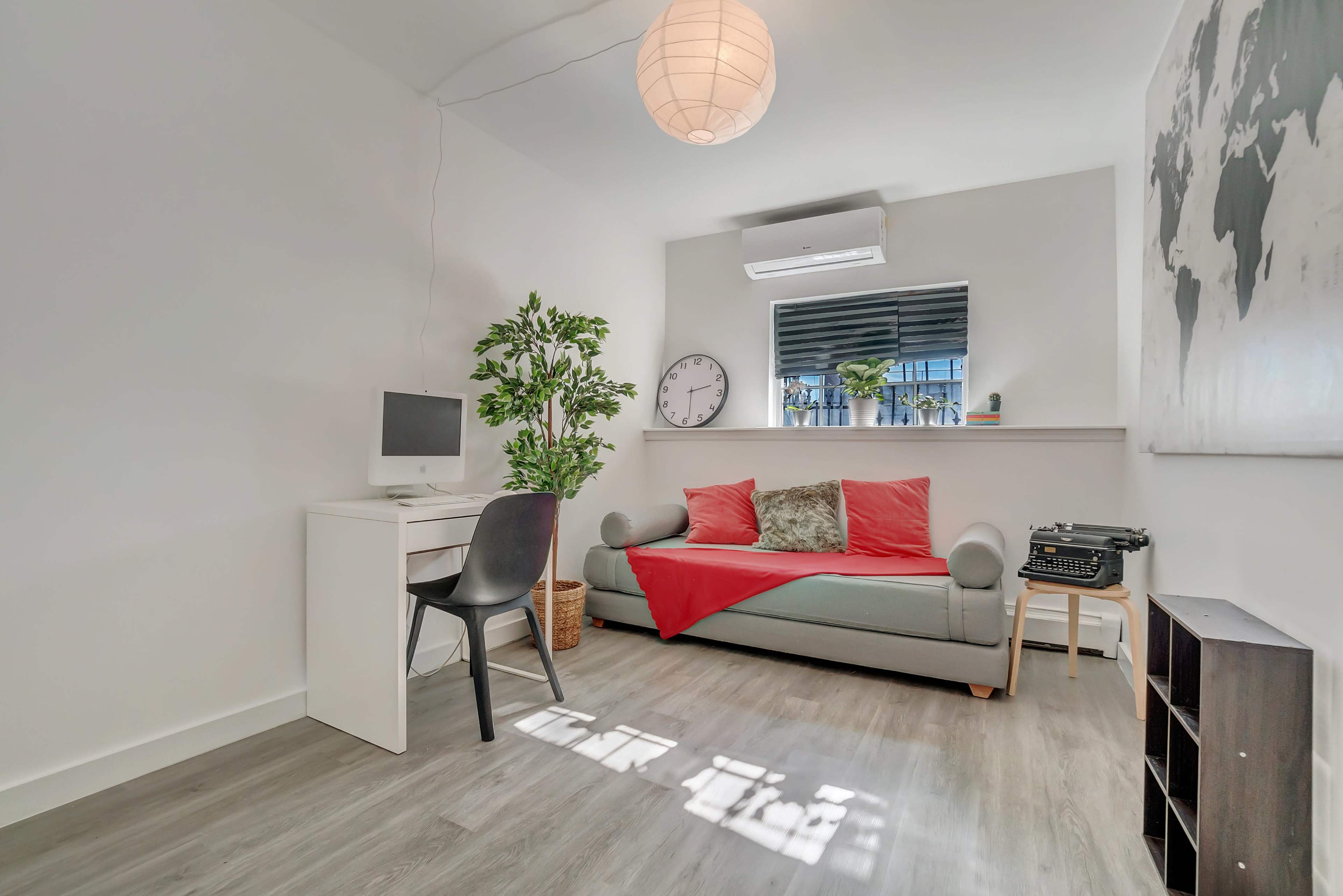 A small, modern room with a gray sofa, a desk with a computer, a clock, a potted plant, and a vintage typewriter.