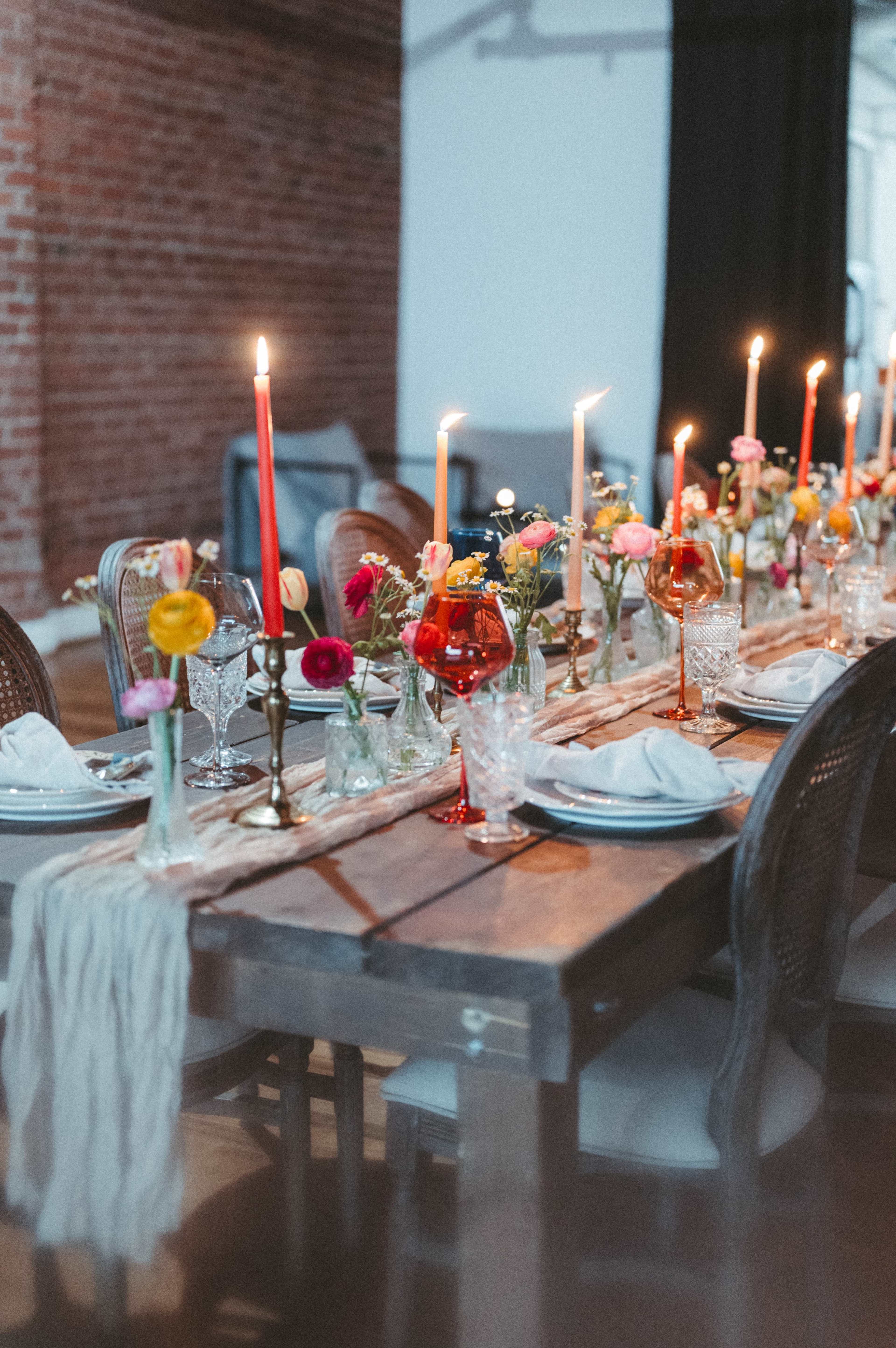 A long wooden dining table is set with various glassware, candles, and colorful flowers in vases, creating an inviting ambiance.
