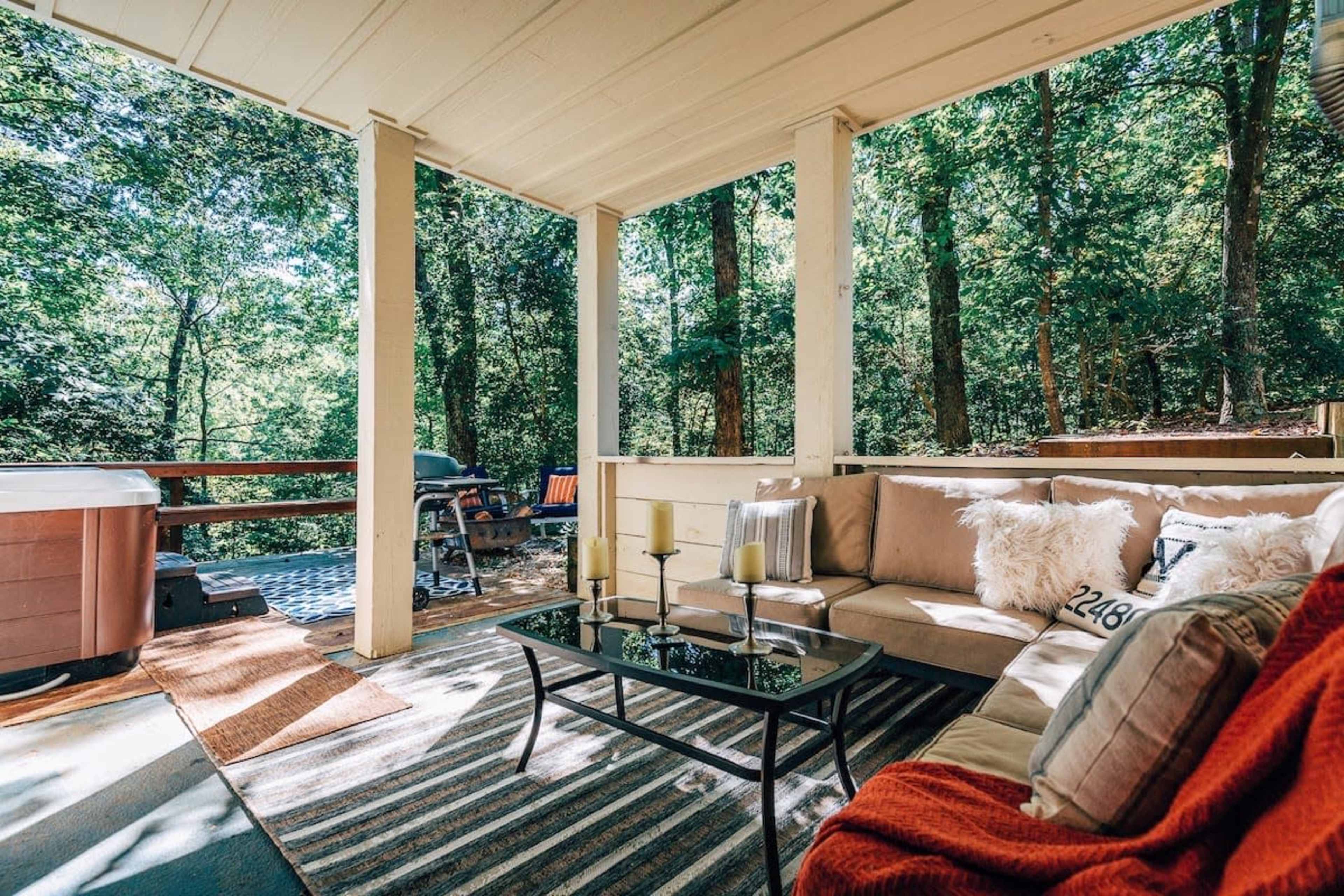 A cozy outdoor seating area features a sectional sofa with cushions, a glass coffee table, and is surrounded by trees with a hot tub visible nearby.