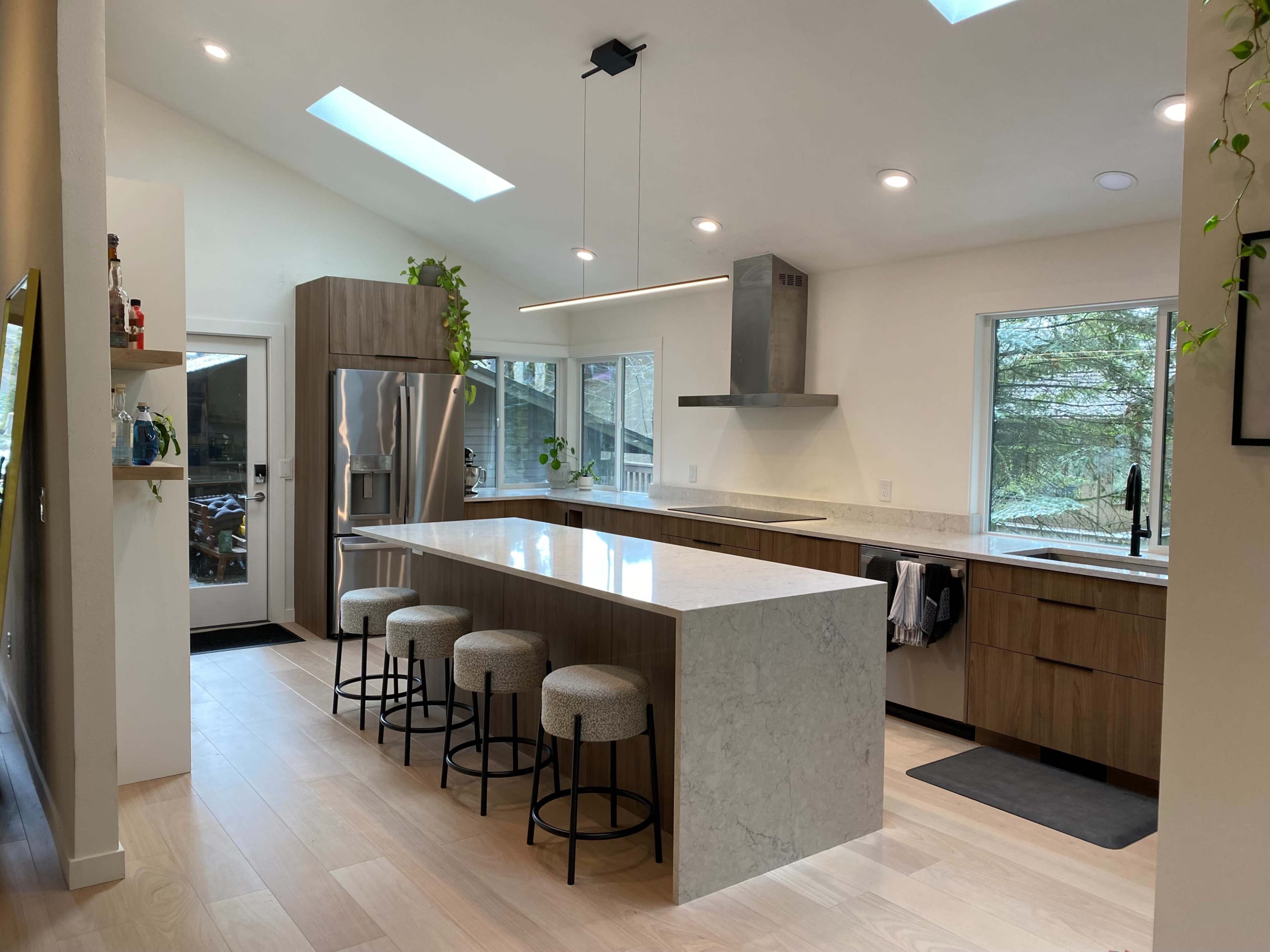 A modern kitchen featuring a large island with three stools, stainless steel appliances, and large windows that provide natural light.