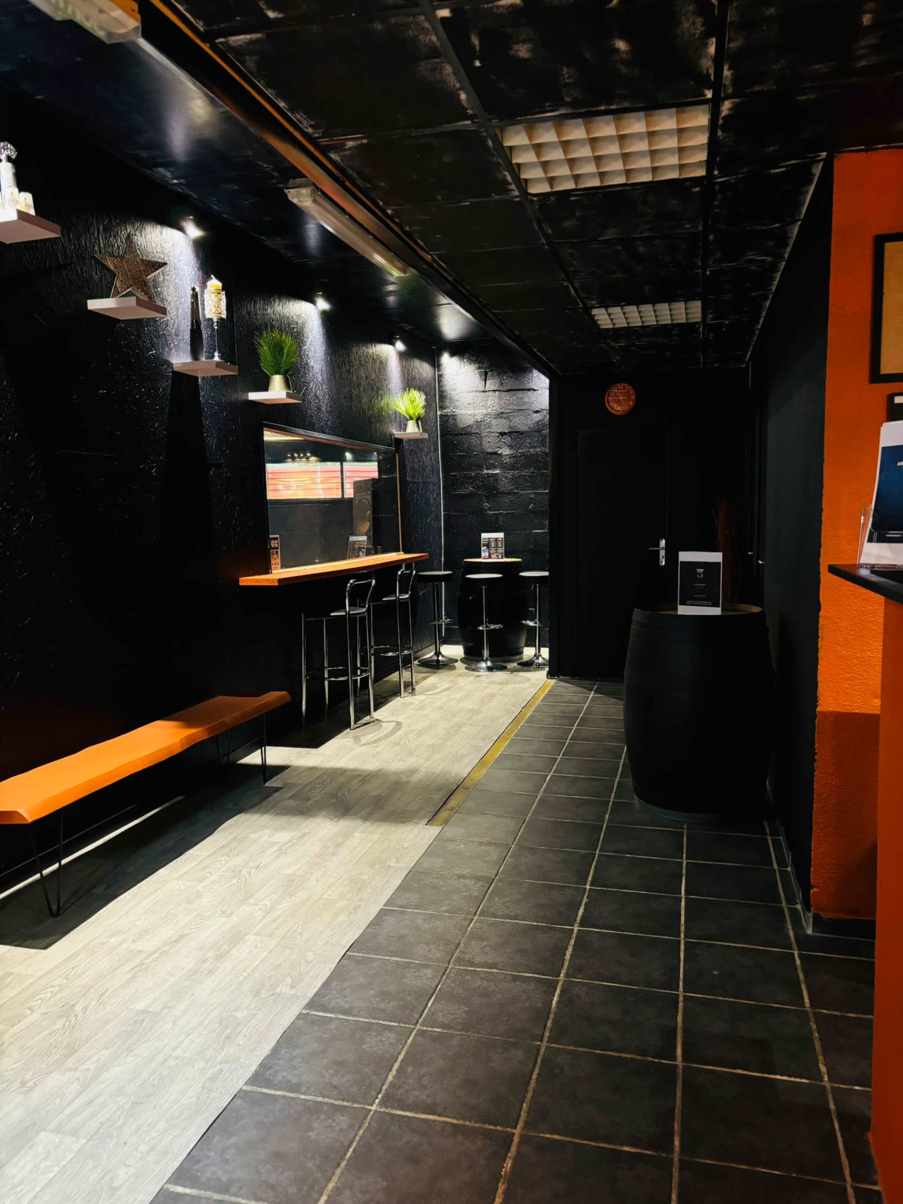 A narrow corridor features dark walls, a bench, and small tables with chairs, illuminated by overhead lights.