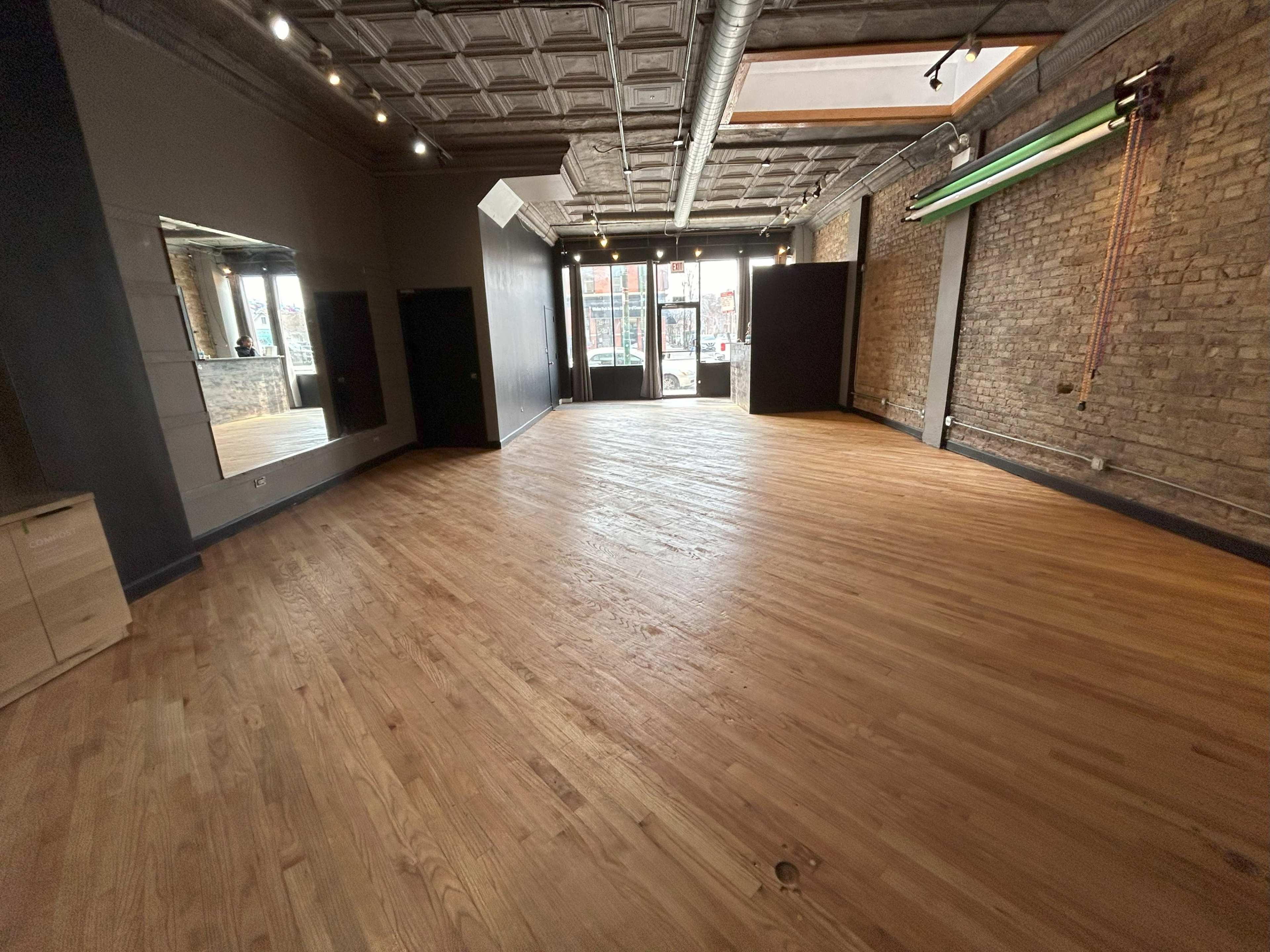 The image shows an empty room with hardwood flooring, exposed brick walls, large windows, and minimal furnishings.