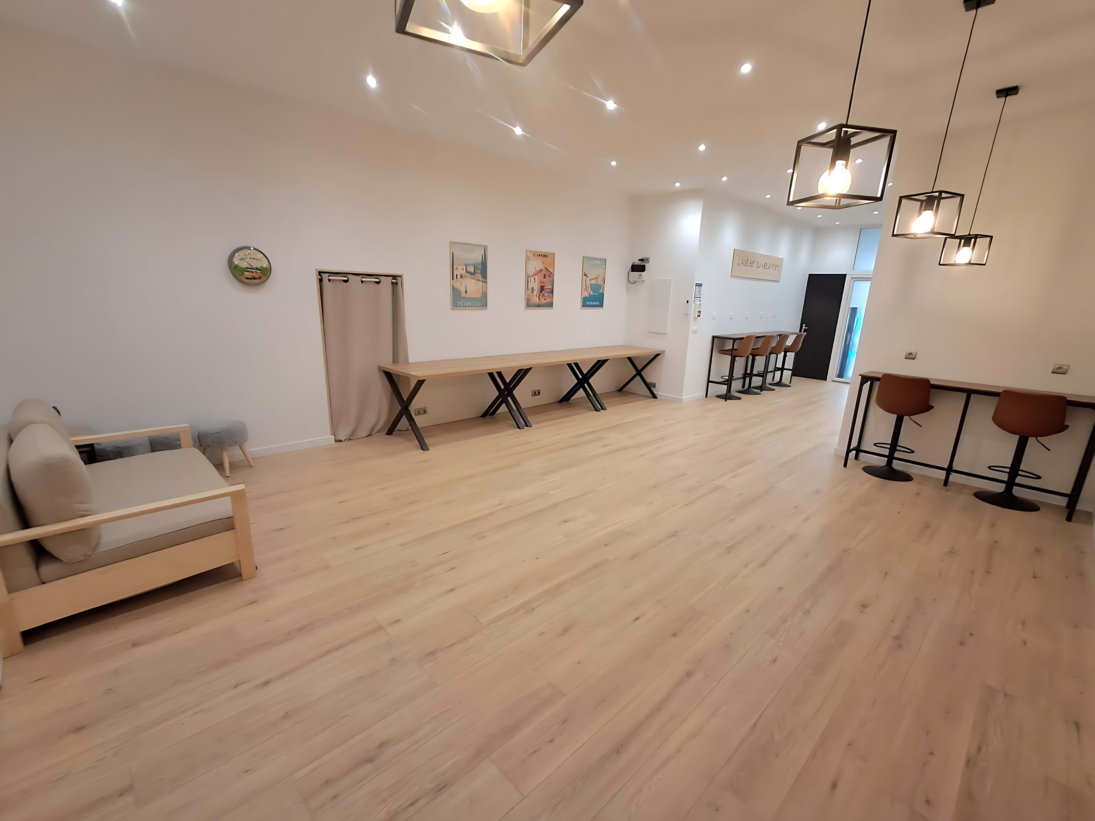 The image shows a spacious, brightly lit interior with wooden flooring, featuring a sofa, two tables, and several bar stools along with wall art.